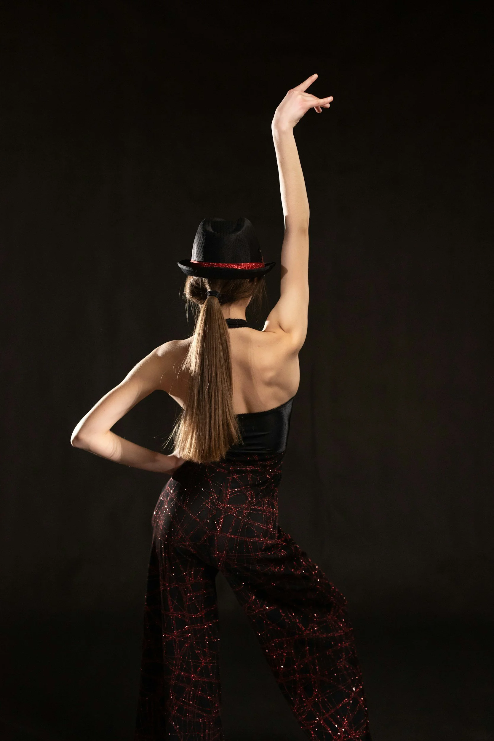 A woman with long brown hair in a ponytail is performing a dance move against a black background. She is wearing a black sleeveless top, red glittery pants with a black netted overlay, and a black hat with a red band.