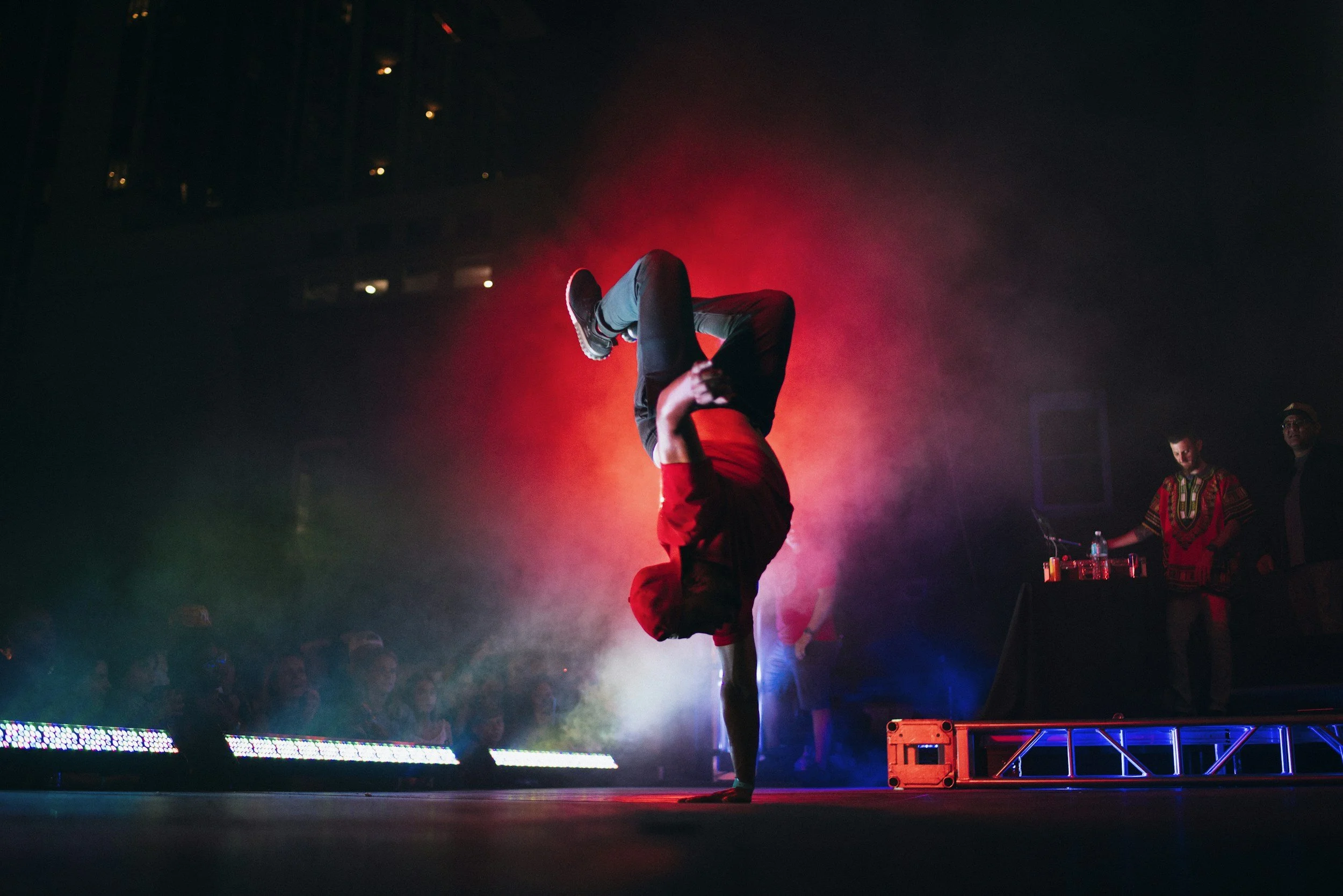 Performer in red outfit balancing upside down on another person's hands on stage with colorful lights and two other individuals in the background.
