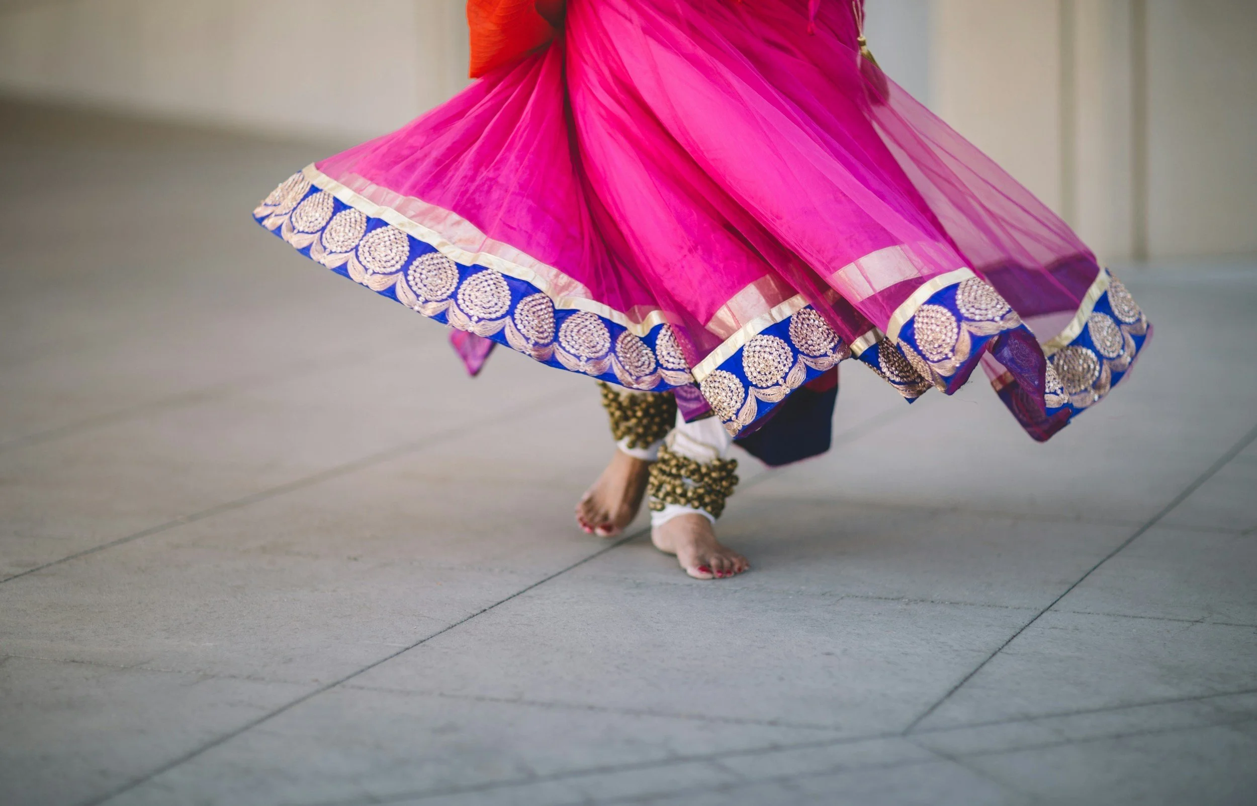 Close-up of a dancer's pink traditional dress with blue and gold embellishments, walking barefoot on a gray tiled floor.