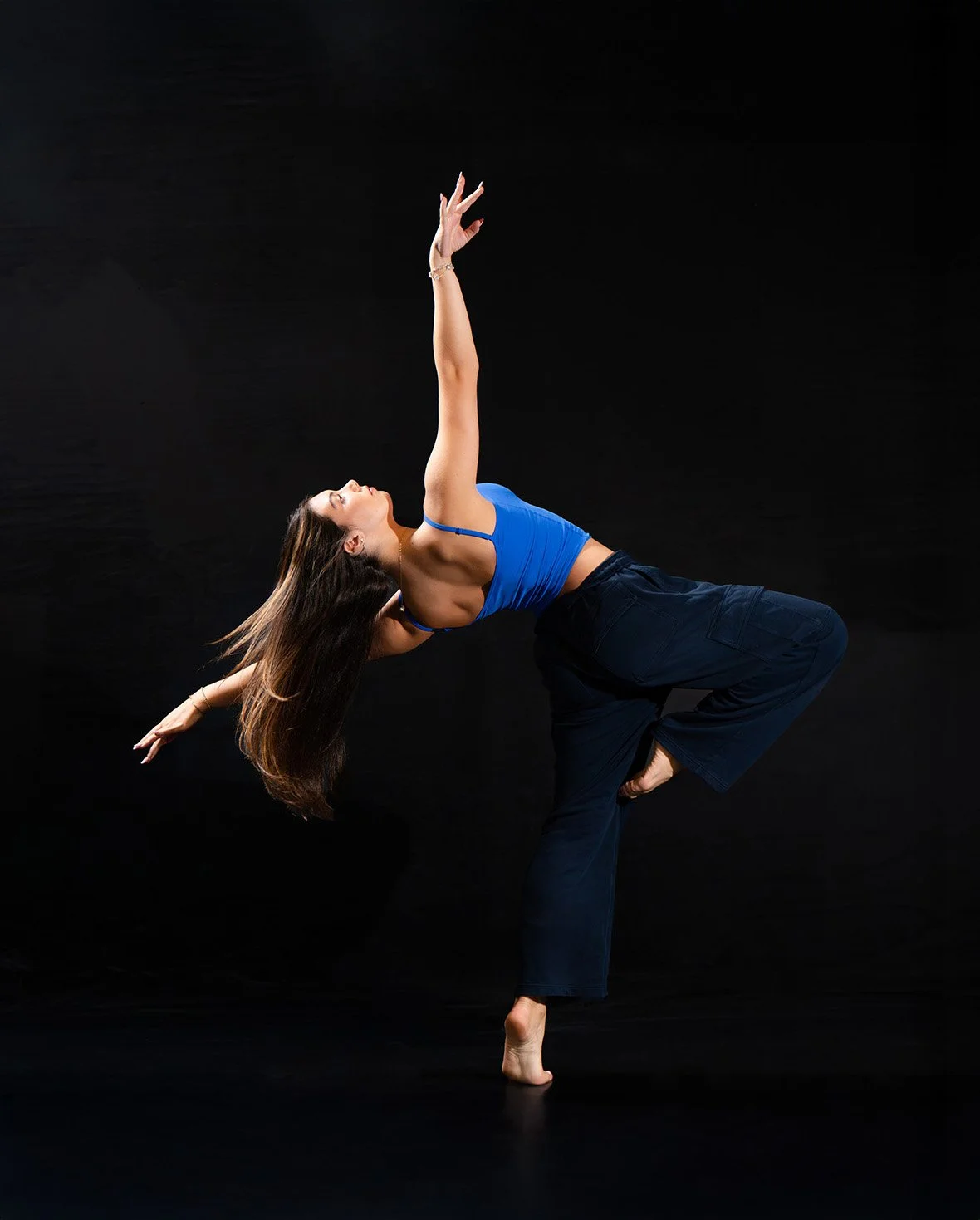 A woman practicing yoga in a pose on a black background, wearing a blue top and black pants.