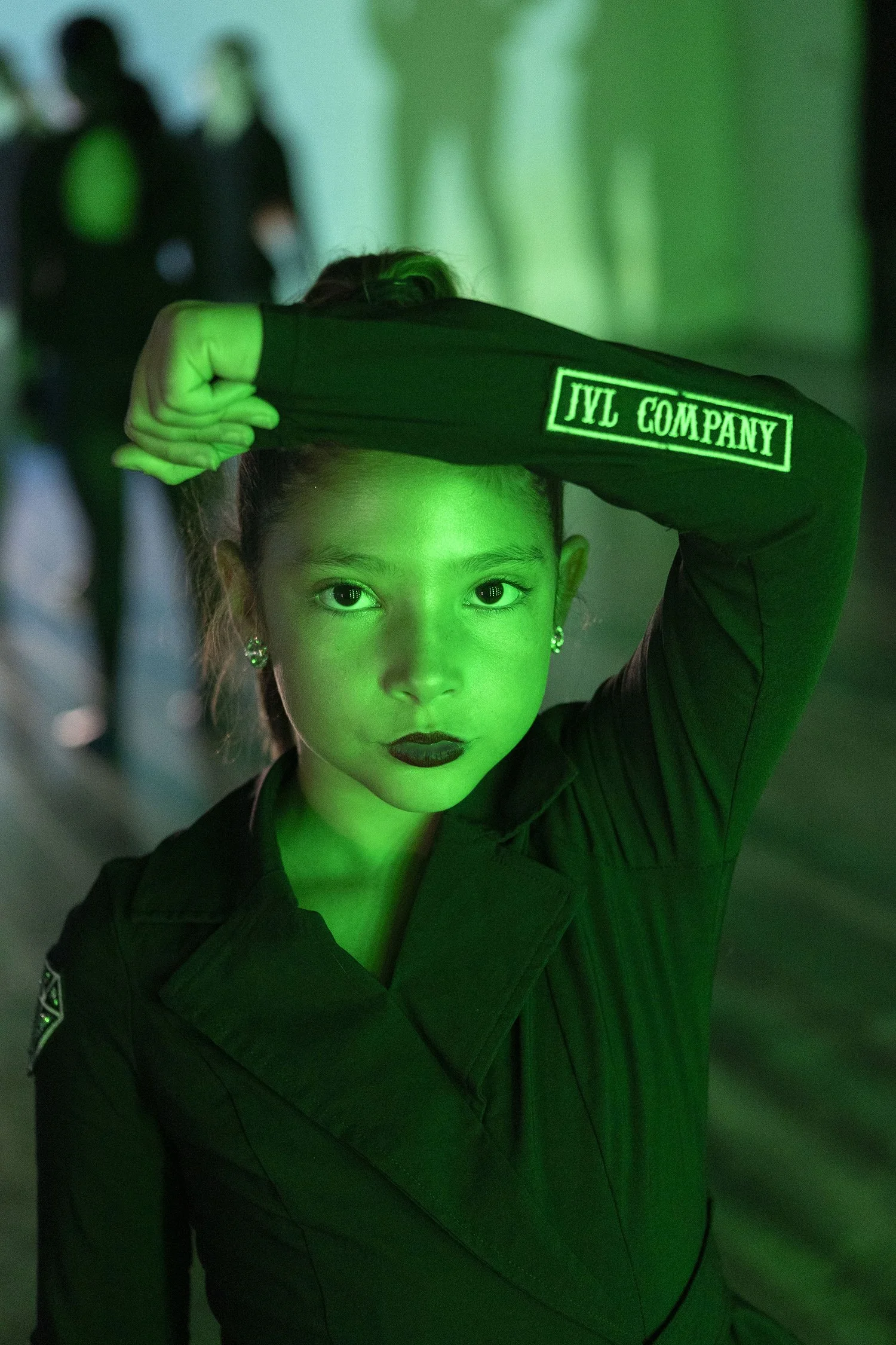A woman with makeup and earrings, wearing a dark uniform with patches, holds her arm up to her forehead, shading her eyes. The scene is illuminated with green light, and there are blurred figures in the background.