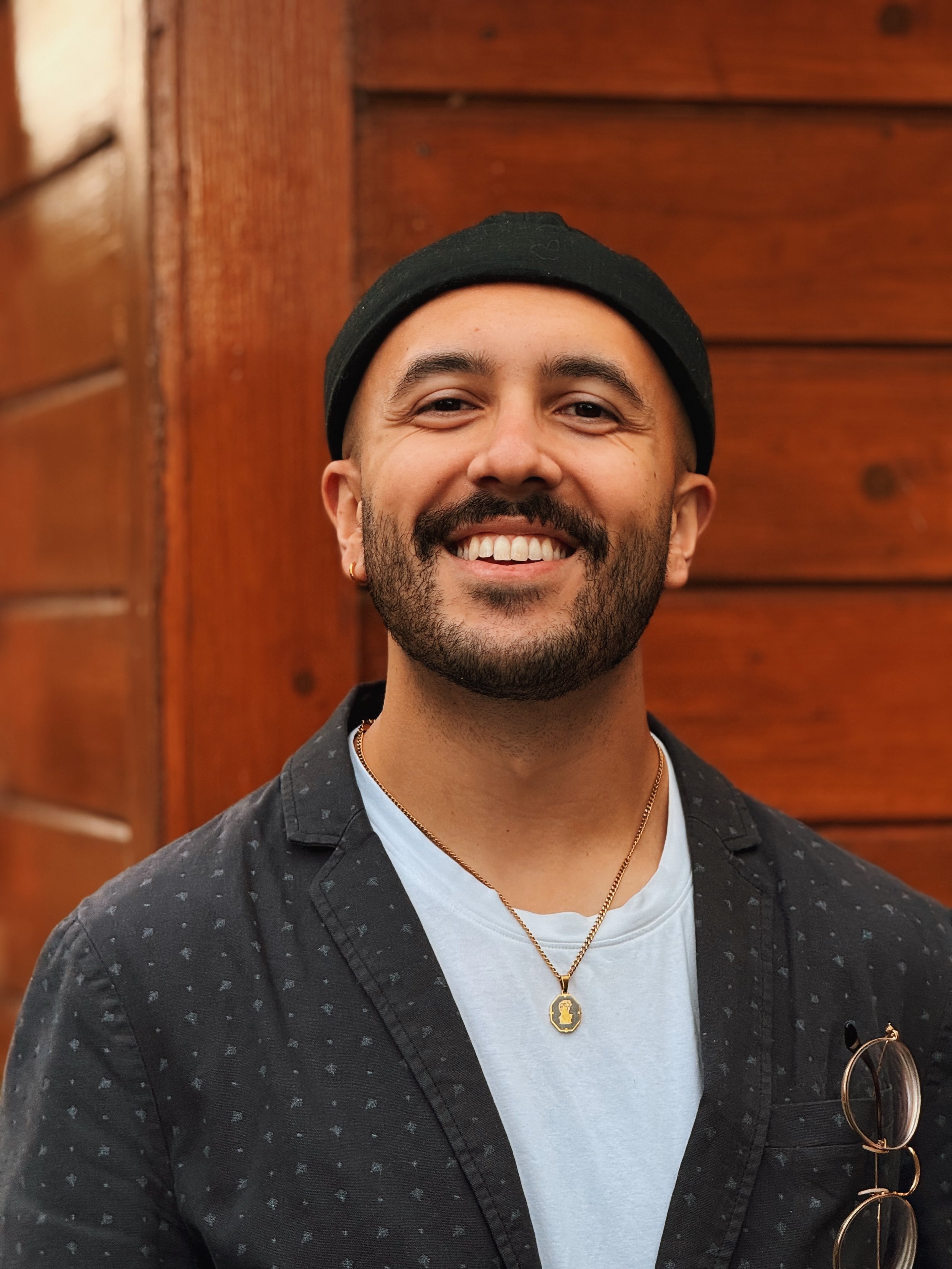 Close-up of a smiling man with facial hair, wearing a black beanie, black patterned shirt, white t-shirt, and gold necklace, standing in front of a wood-paneled background.