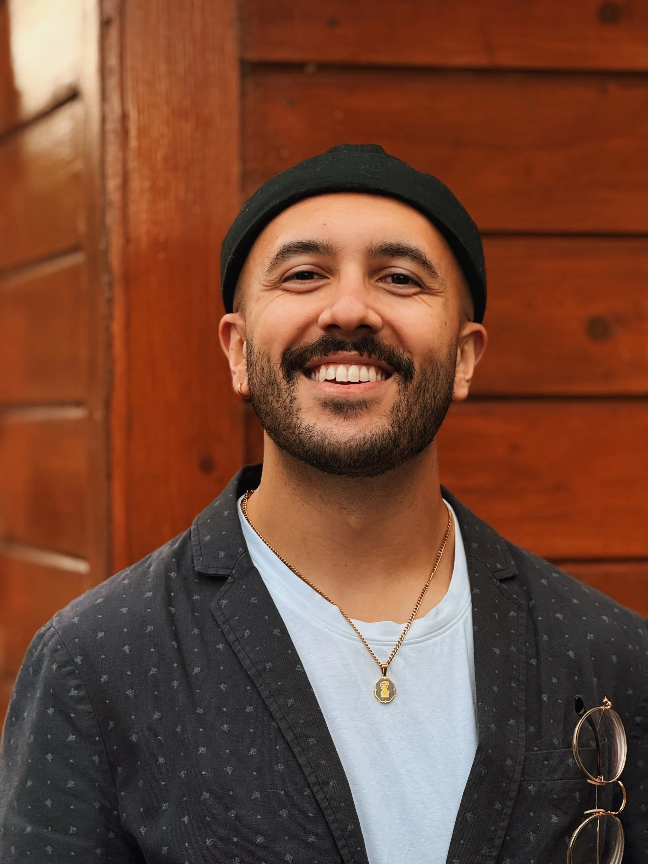 Close-up of a smiling man with facial hair, wearing a black beanie, black patterned shirt, white t-shirt, and gold necklace, standing in front of a wood-paneled background.