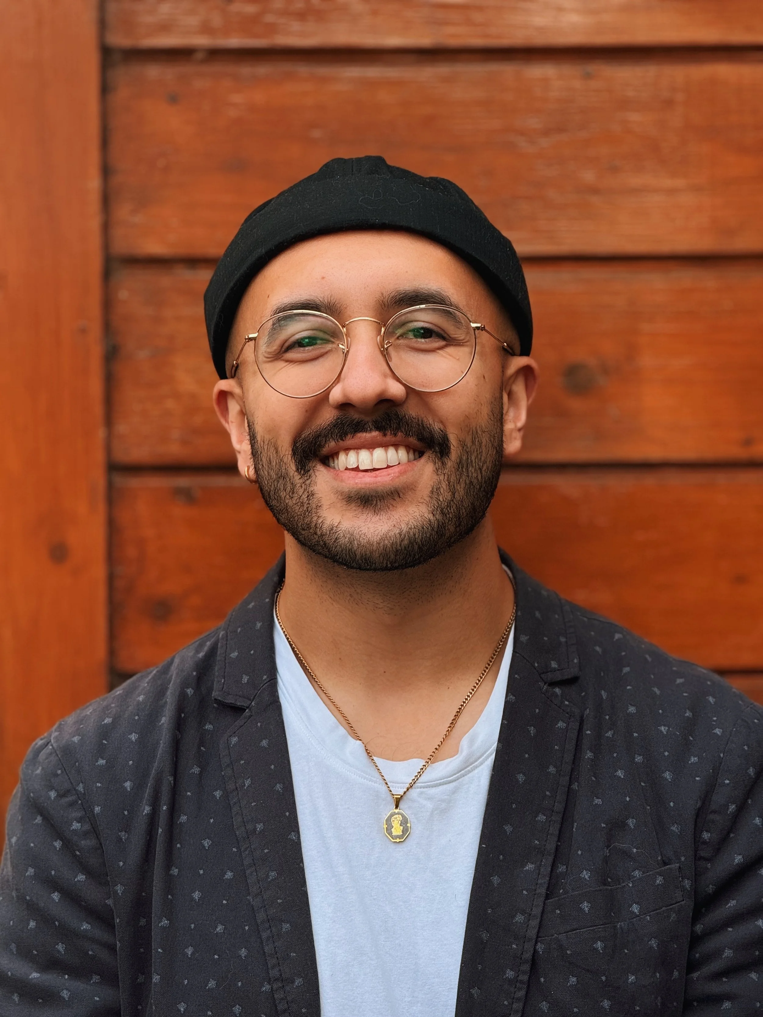 A smiling man with glasses, a black beanie, a black patterned jacket, a white T-shirt, and a gold necklace with a pendant, standing in front of a wooden wall.