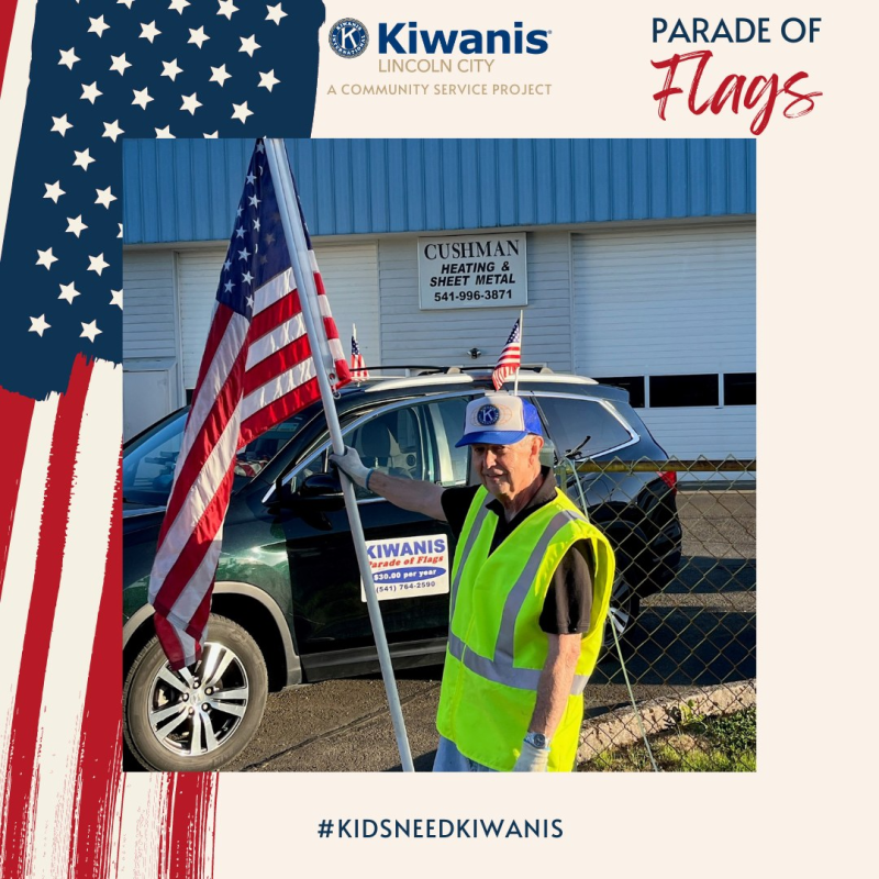 Pat "Fun Pa" Dunn wearing a blue Kiwanis hat and a yellow safety vest holding an American flag during a parade of flags event in Lincoln City, standing next to a black vehicle with a Kiwanis banner.