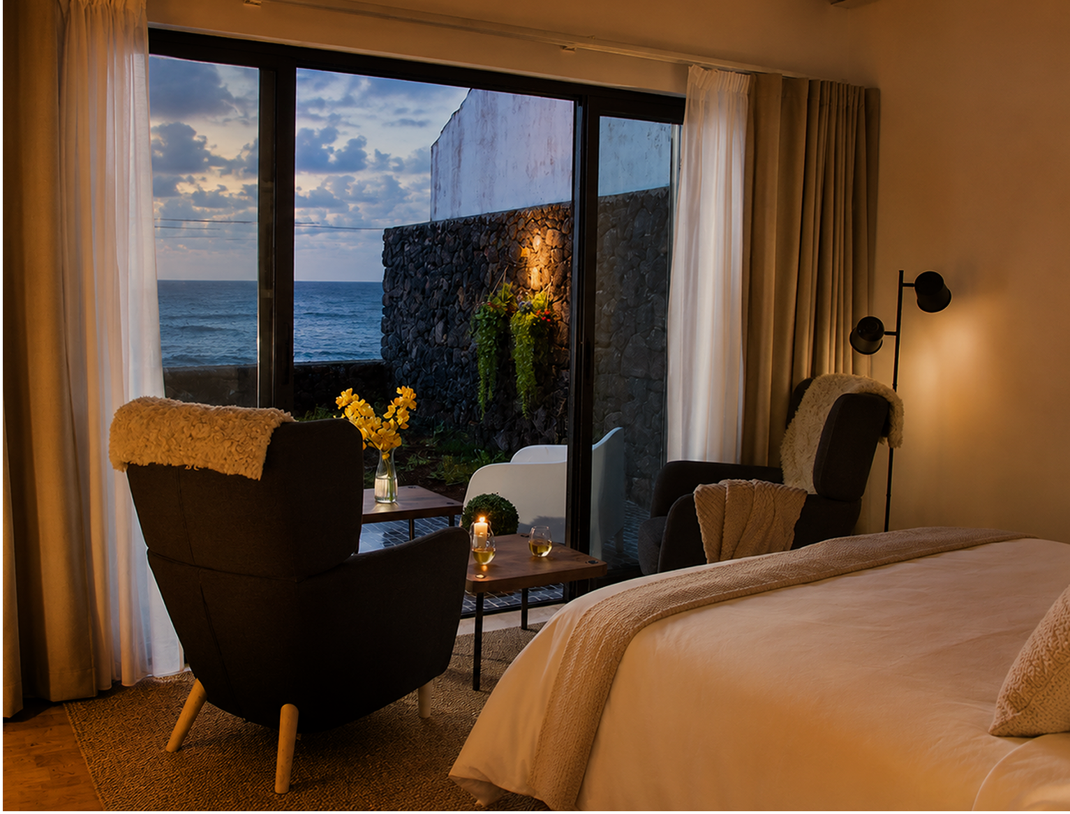 Hotel room with large glass sliding door overlooking the ocean during sunset, decorated with chairs, a small table with a candle and glasses, and indoor plants.