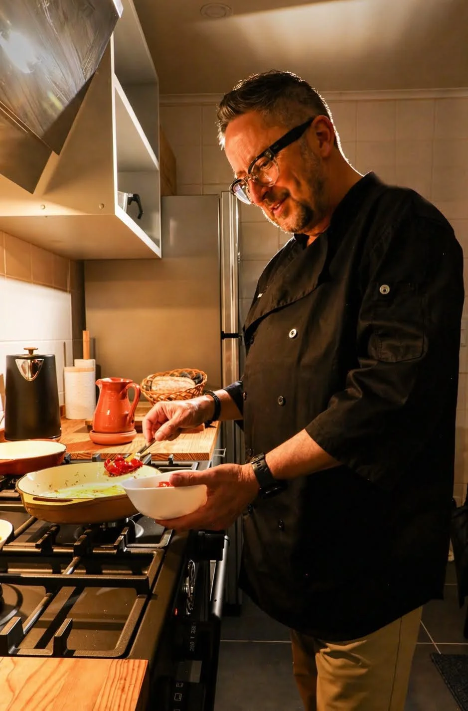 A man in a black chef's coat and glasses cooking in a kitchen, holding a white bowl and a spoon, with various kitchen items around him.