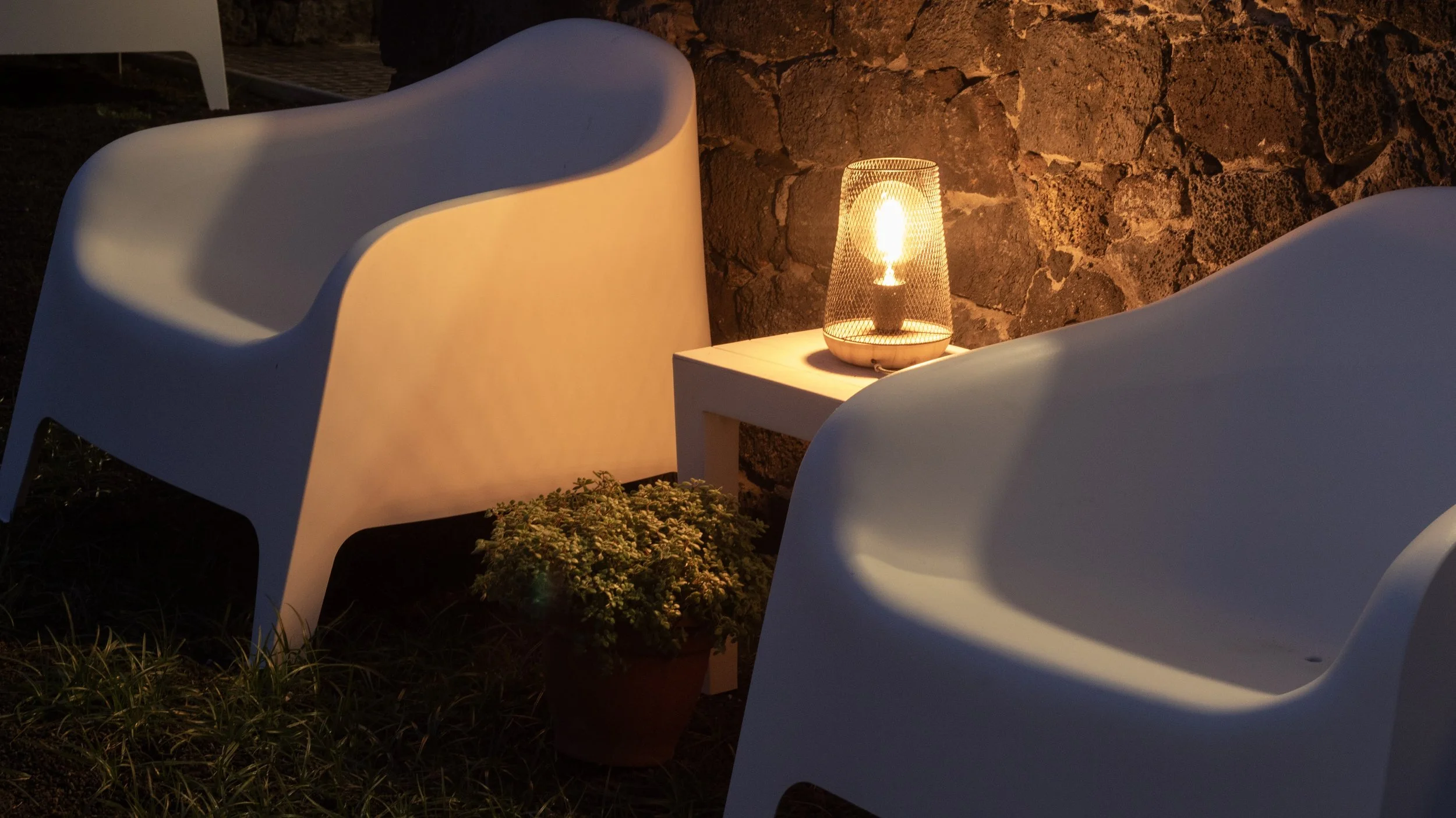 Two white chairs and a small table with a lit decorative lamp on it, situated outdoors against a stone wall.