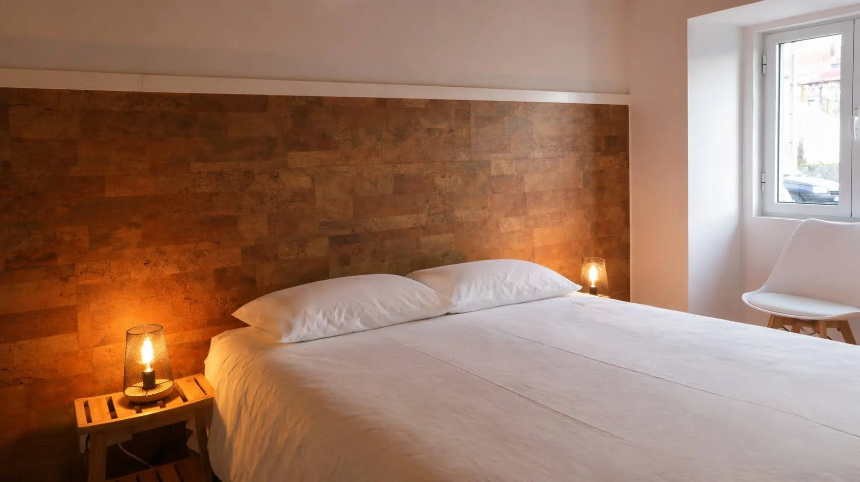 A bedroom with a large bed featuring white linens and pillows, a headboard with a wood texture, two small bedside lamps, a window with natural light, and a white chair near the window.