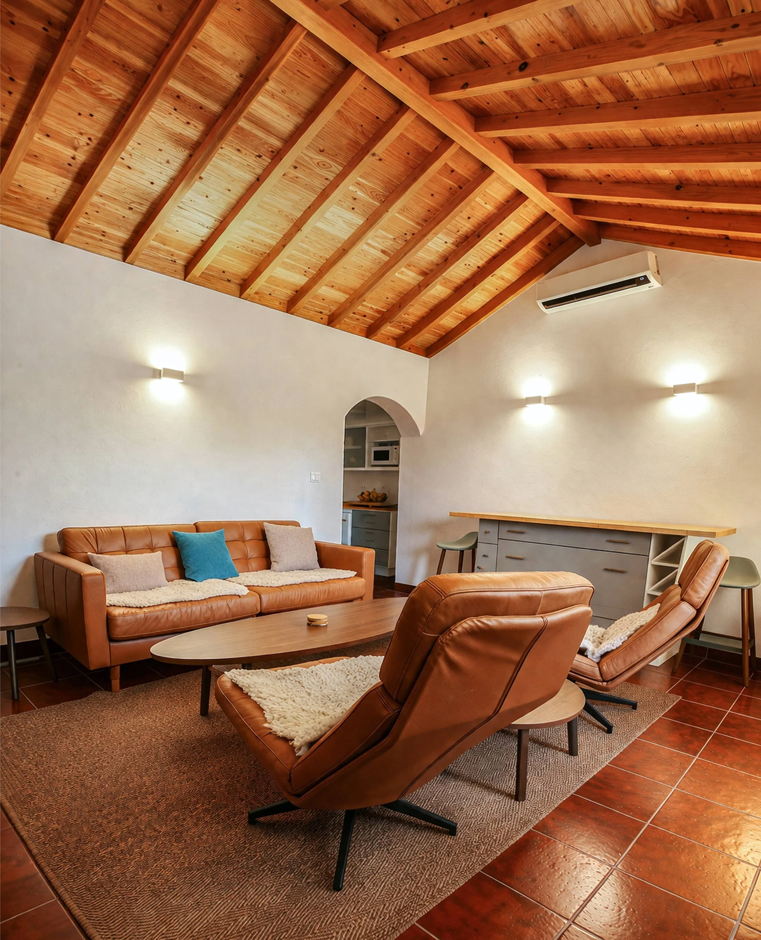 Living room with brown leather furniture, a wood coffee table, a large patterned rug, a wooden ceiling with beams, and white walls with wall-mounted lights.