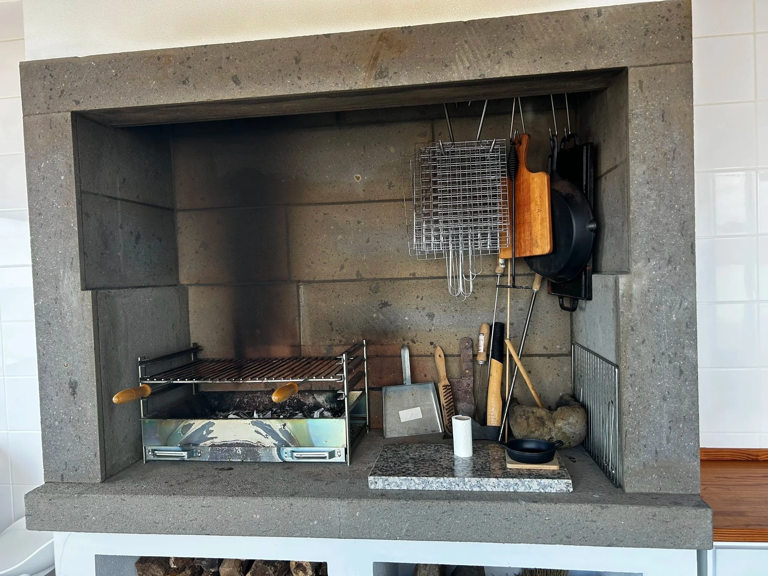 A fireplace with a kitchen grill inside. The fireplace is surrounded by a gray stone mantel and has a fire grate with a grill set inside. Various tools are hanging or placed around, including a metal wire rack, a wooden handled fork, a pan, a paper t