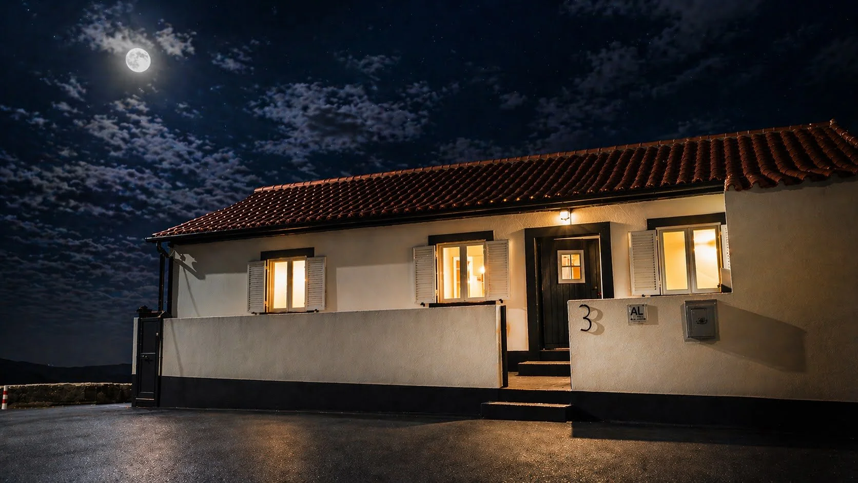 A house under a cloudy night sky with a bright full moon. The house has a red tiled roof, white walls, and several illuminated windows.