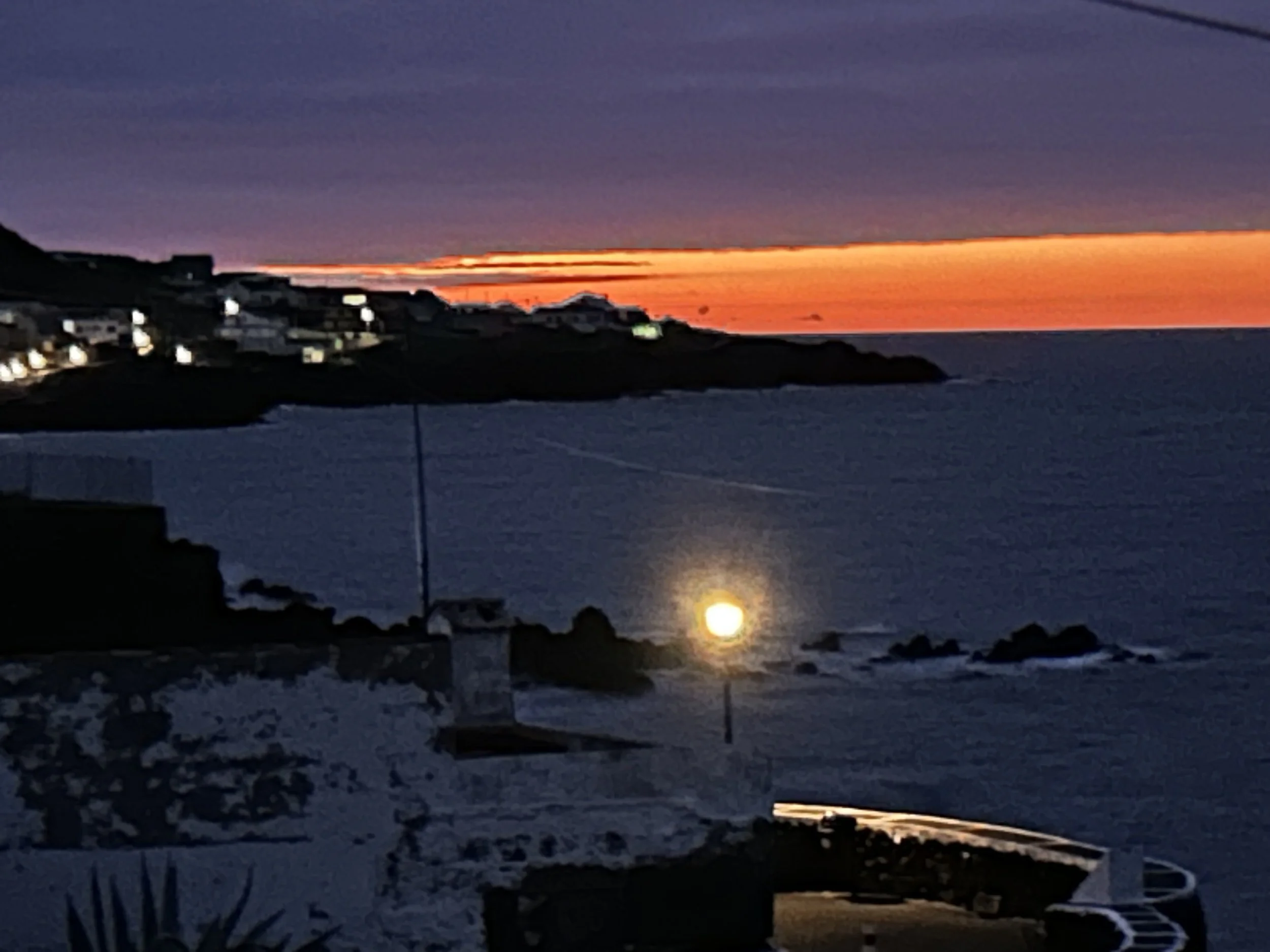 A sunset over the ocean with orange and purple hues and a silhouette of a hillside with houses on the left.