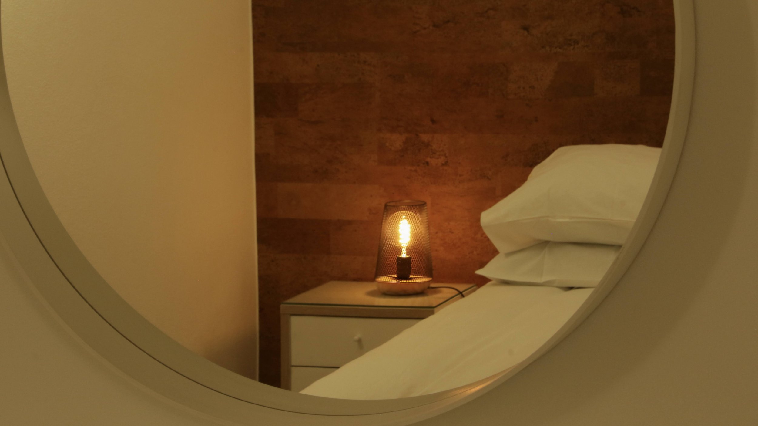 A bedside table with a glowing lamp and pillows, viewed through a mirror.