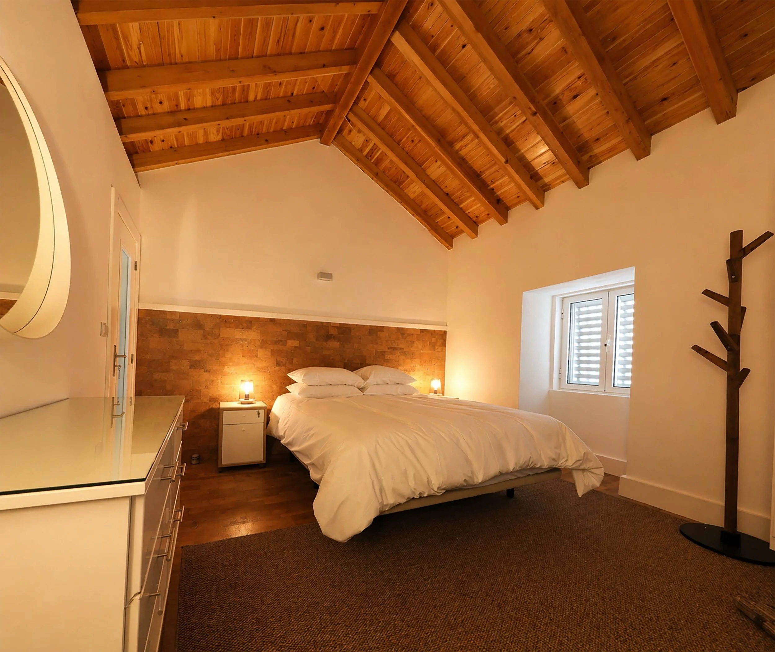 A cozy bedroom featuring a wood-paneled ceiling with exposed beams, a bed with white sheets and pillows, a brick accent wall behind the bed, a small white nightstand with a lamp, a window with white shutters letting in natural light, and a dark brown
