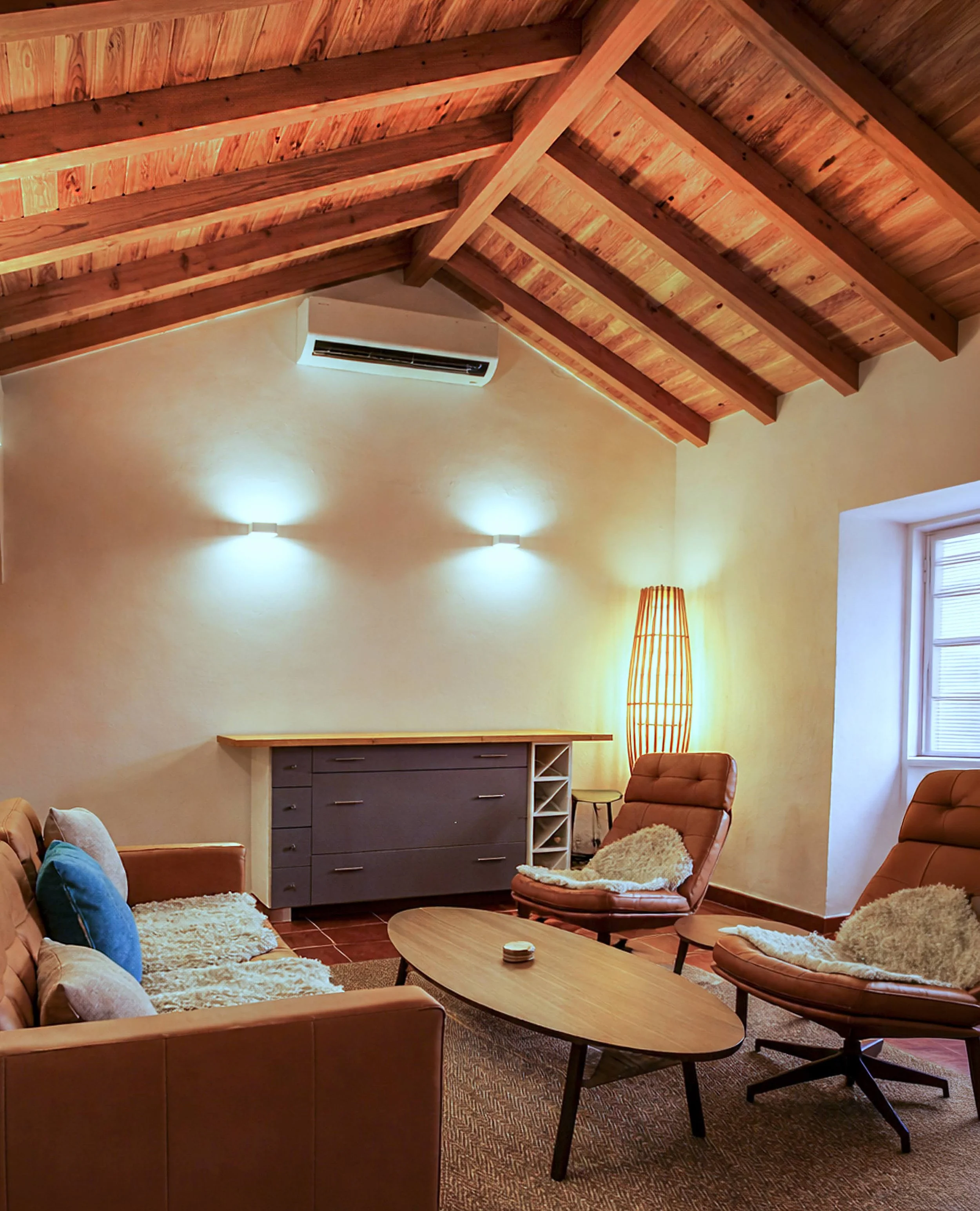 Living room with a wooden vaulted ceiling, beige walls, a beige sofa with blue and beige pillows, a brown leather armchair with a sheepskin throw, a wooden coffee table, a tall wicker floor lamp, and a window with blinds.