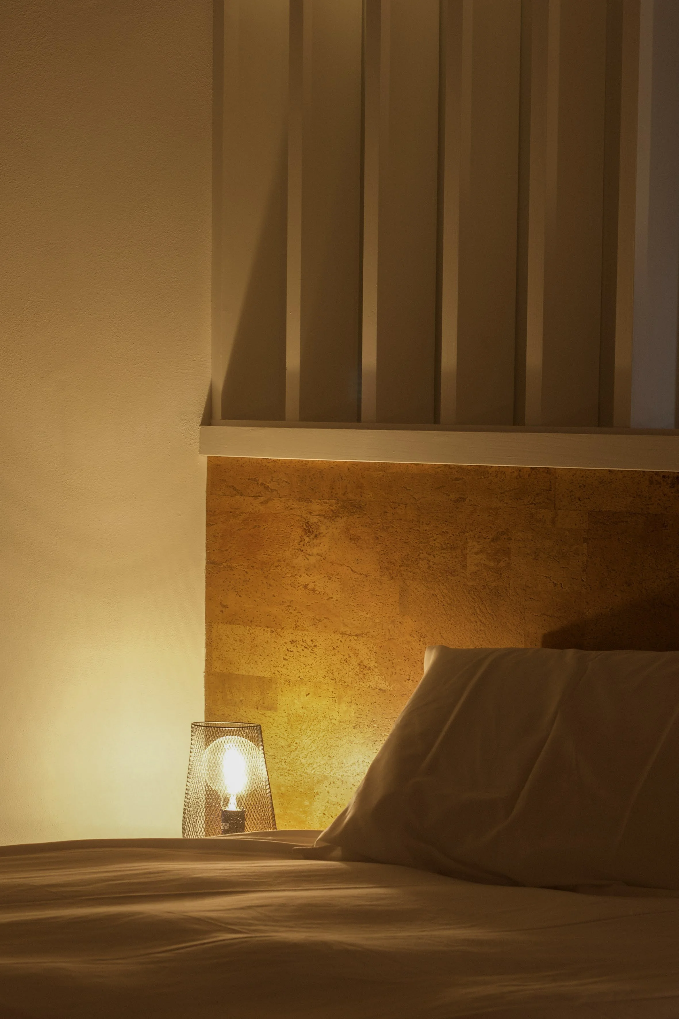 A cozy bedroom with a bed, a bedside lamp, and a textured wall in warm lighting, featuring a wooden headboard and a ceiling with wooden slats.