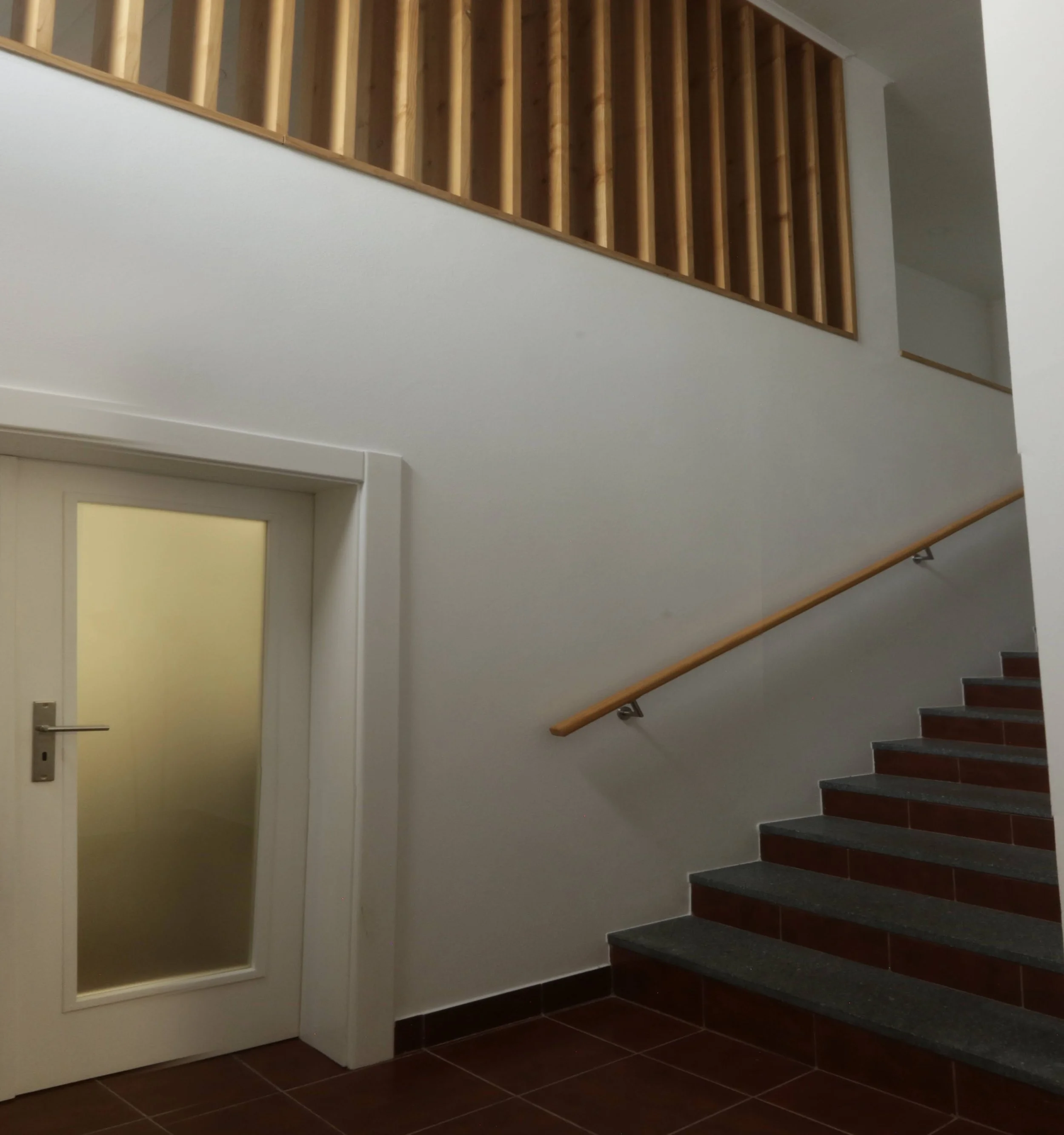 Interior view of a staircase with dark steps, a wooden handrail, and a railing with vertical wooden balusters. There is a white wall with a door and door frame.