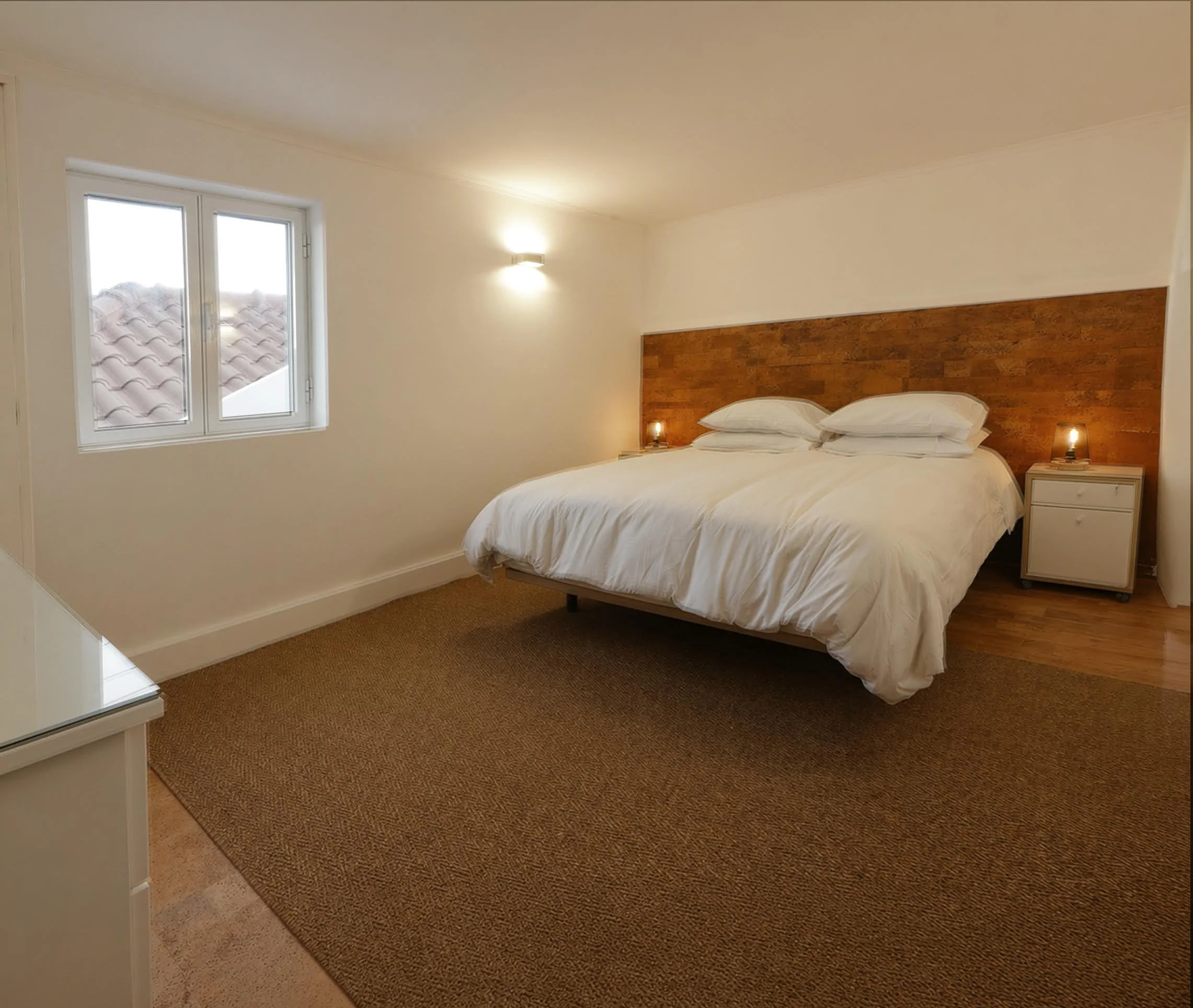 A bedroom with a bed that has white bedding and pillows, a wooden headboard, two bedside tables with lamps, a window with two sections showing a tiled roof outside, beige walls, and a brown carpeted floor.