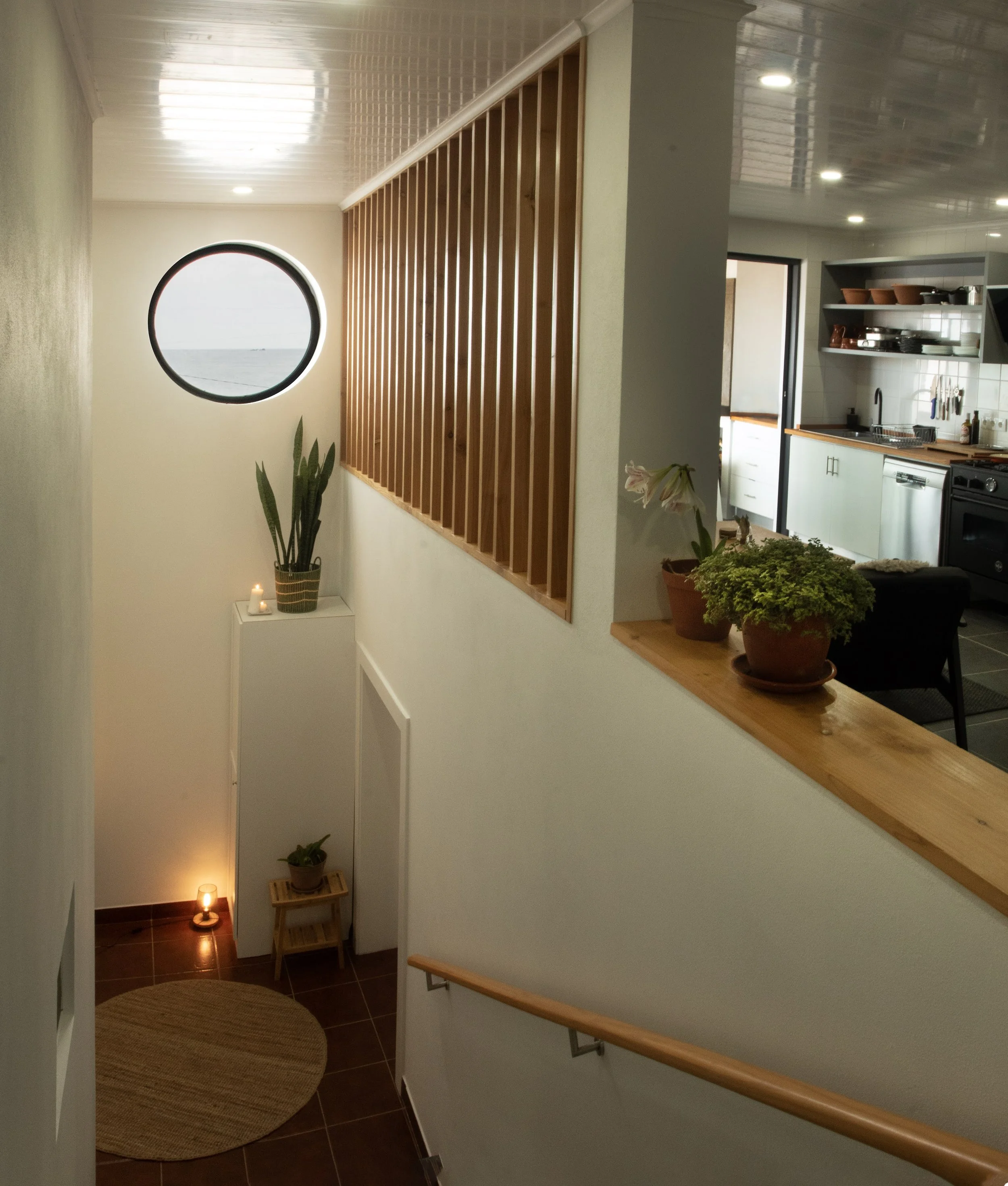 Interior view of a modern home with a staircase, potted plants, candles, and a large circular window showing a view of water and sky.