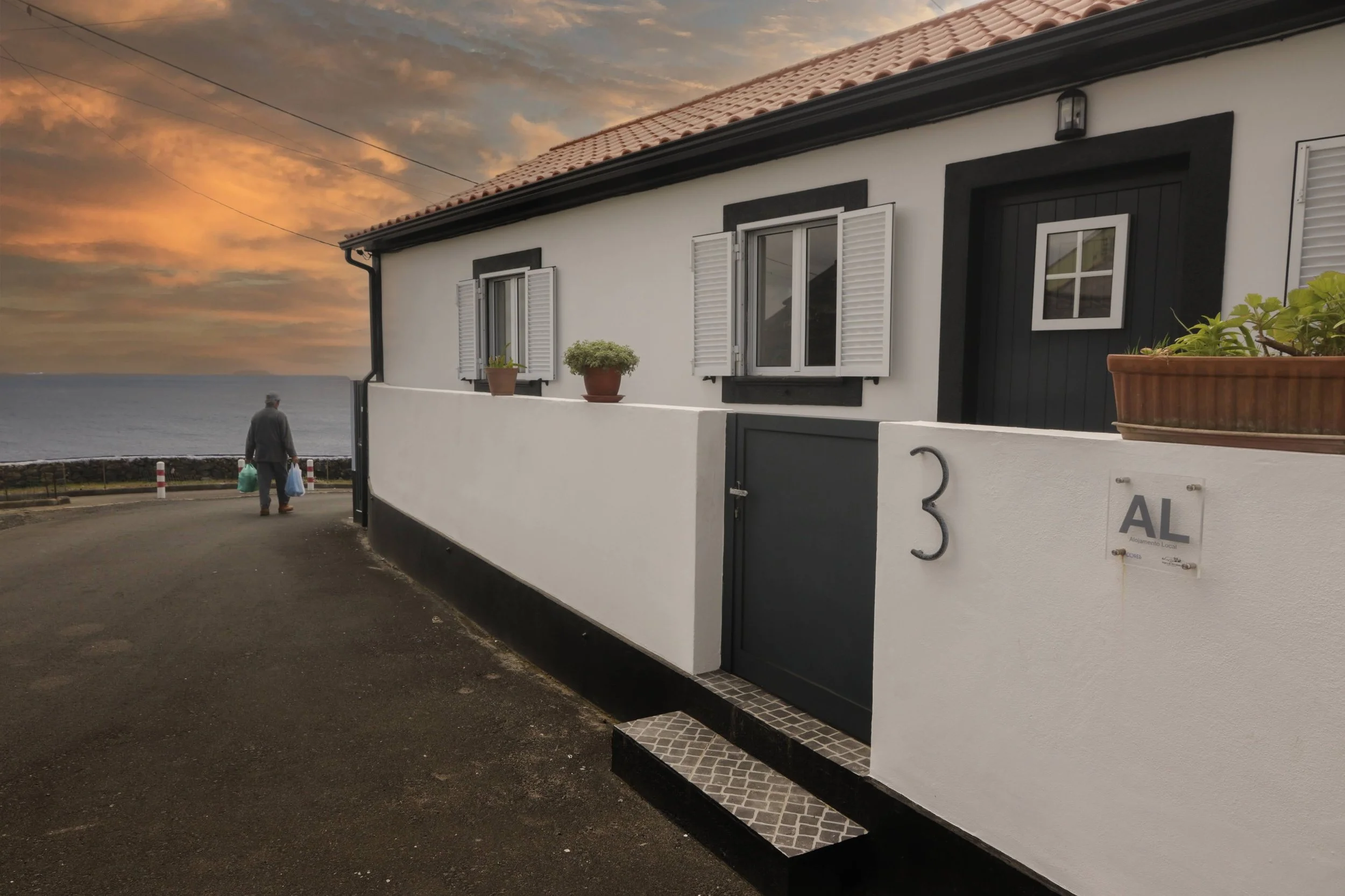 A white house with black trim and shutters, two potted plants on the ledge, a person walking away carrying bags, near the ocean at sunset.