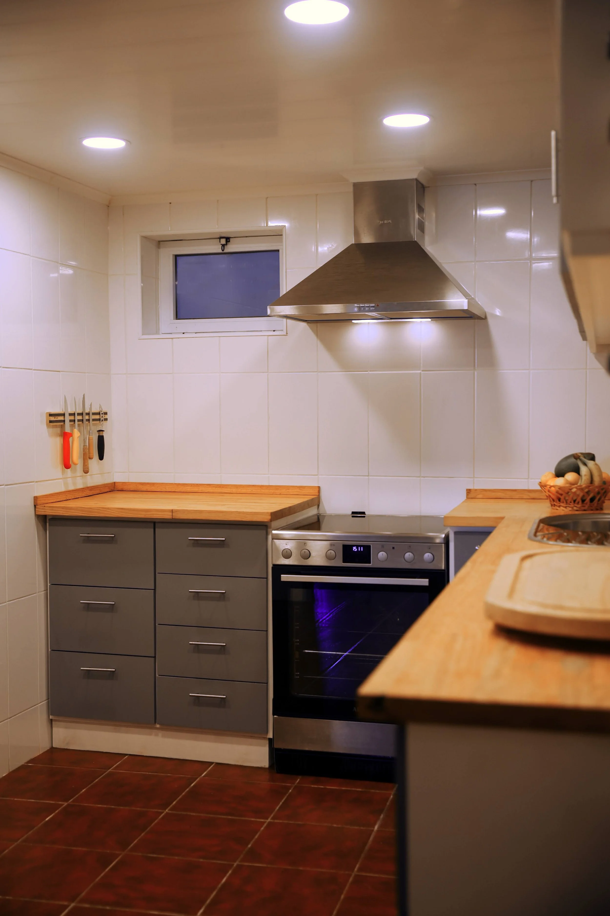 A modern kitchen with white tiled walls, a wooden countertop, gray cabinets, a stainless steel stove and range hood, a small window, and a basket of bananas and oranges on the countertop.