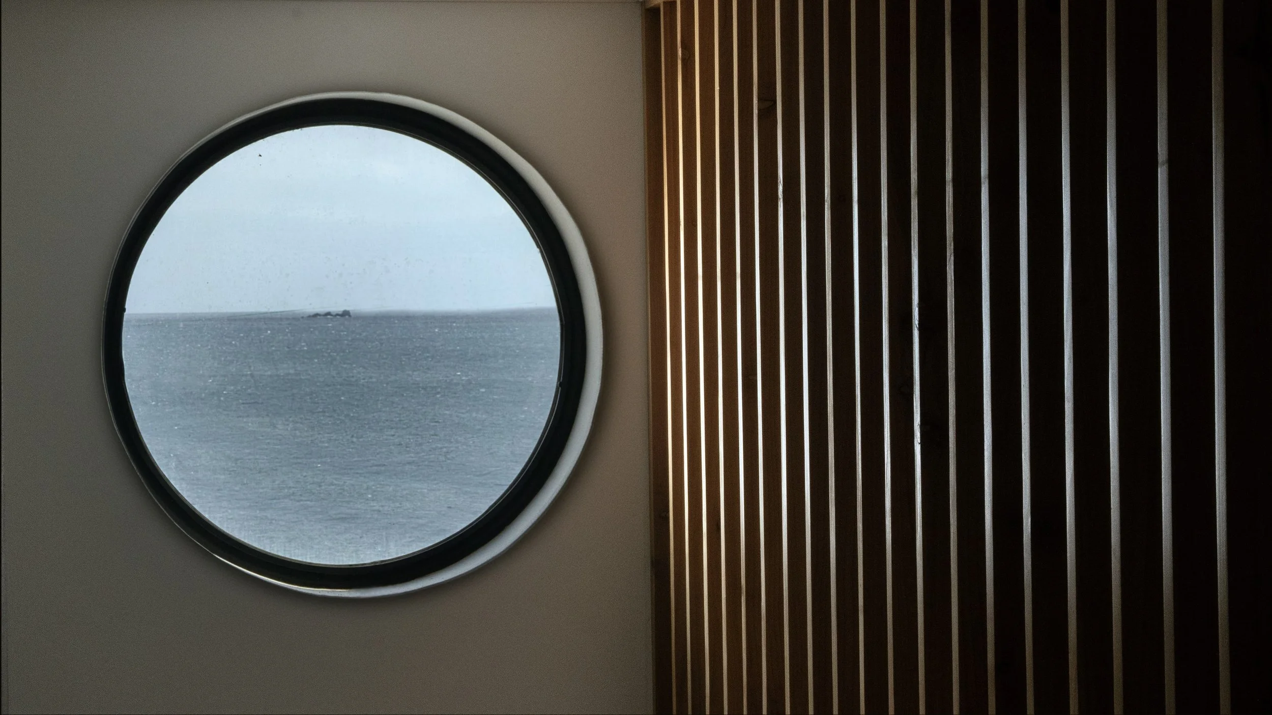 View through a circular window showing a body of water and a small boat in the distance, with wood paneling on the right side.