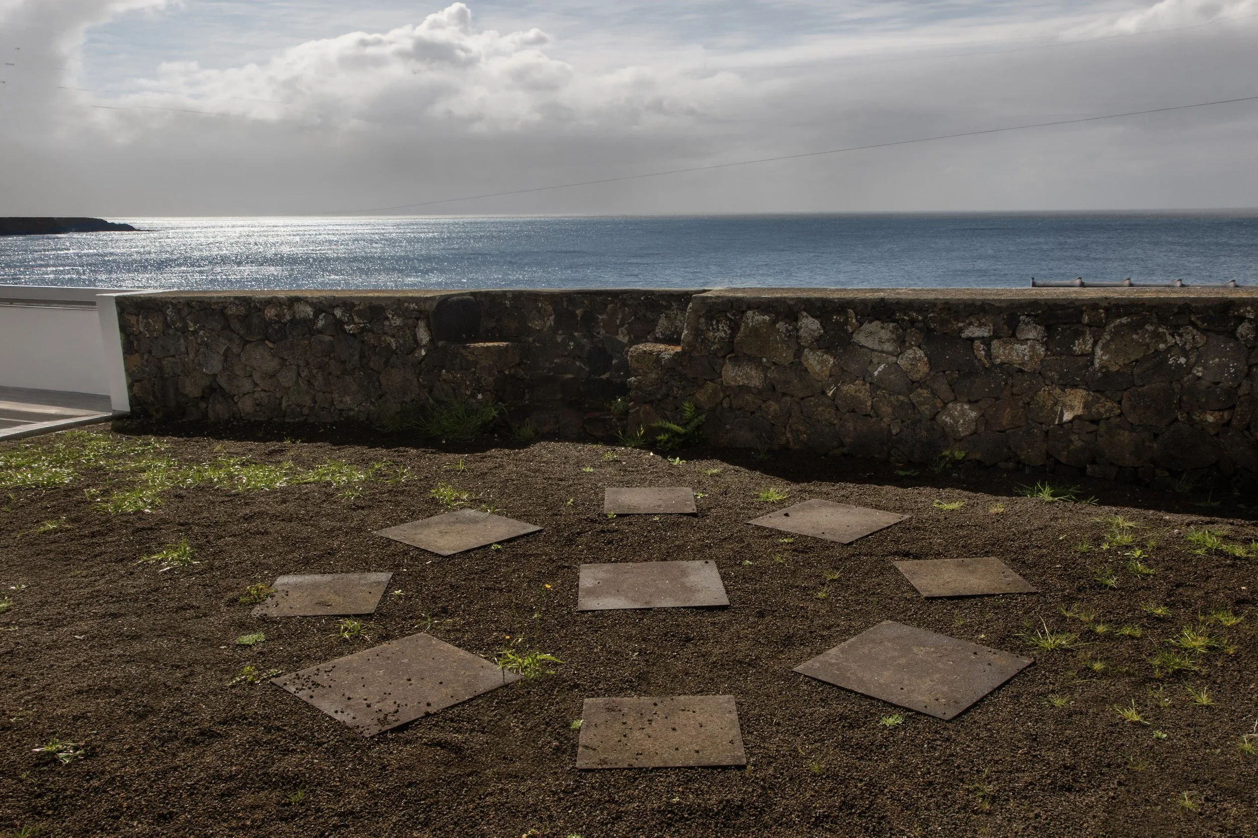 A stone wall separates a dirt area with square stepping stones leading toward the ocean. The ocean is calm with sunlight reflecting off the water, under a partly cloudy sky.