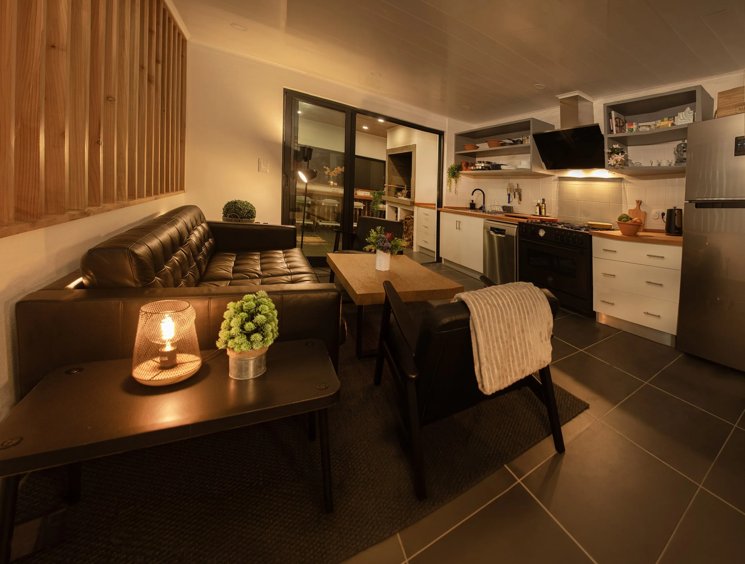 A modern kitchen and living room combination with a dark leather sofa, a small table with a floral arrangement, and open shelving with kitchen essentials. Large sliding glass doors lead to an outdoor patio.