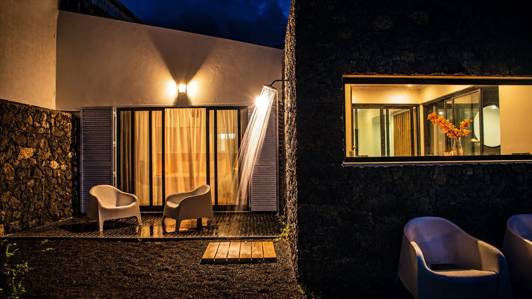 Nighttime outdoor patio area with two chairs, a waterfall feature, and a window showing a room with a vase of orange flowers, black stone walls, and warm interior lighting.