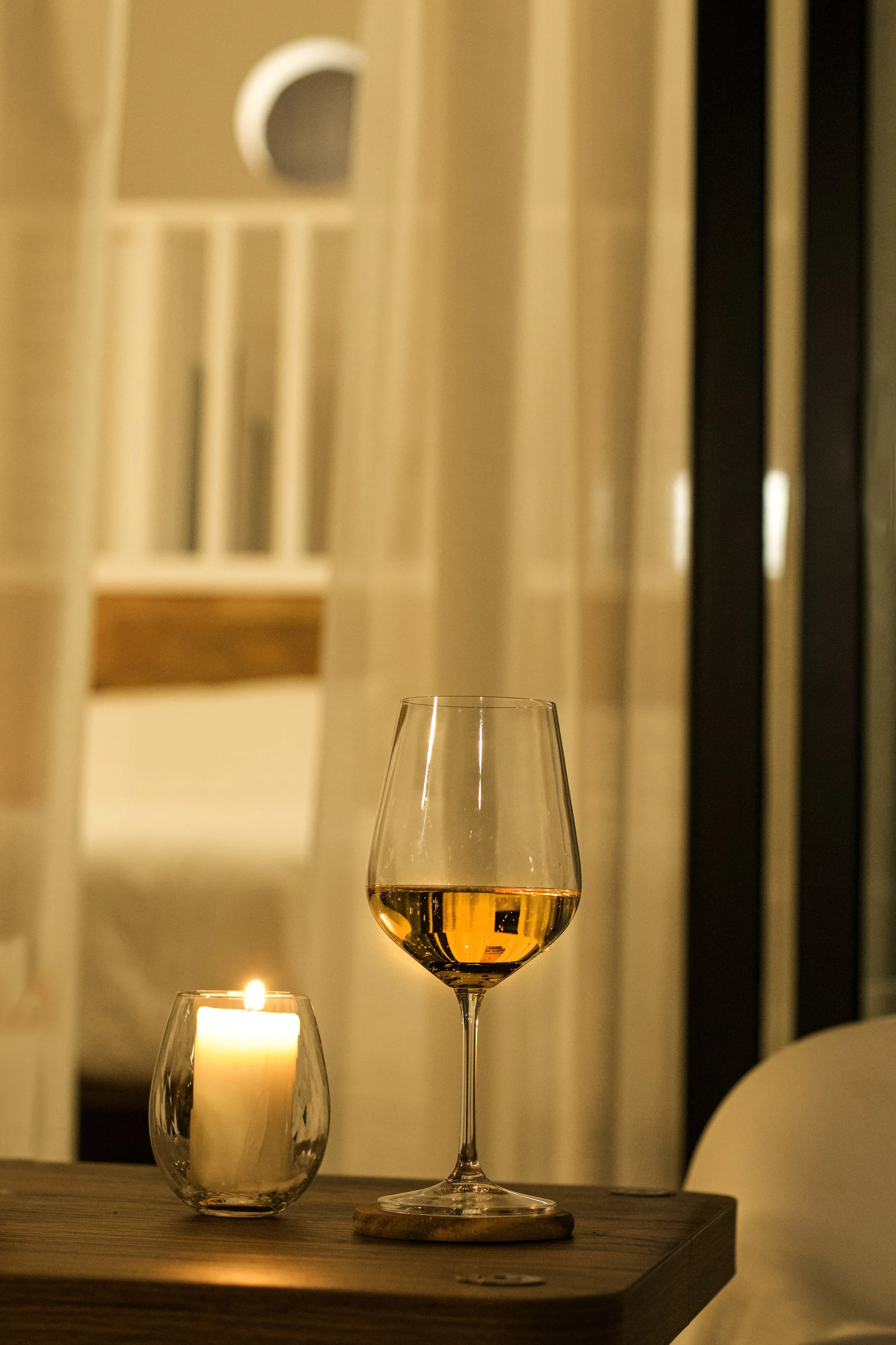 A glass of white wine on a wooden table, a candle in a glass holder, and a bar with a curtain in the background.