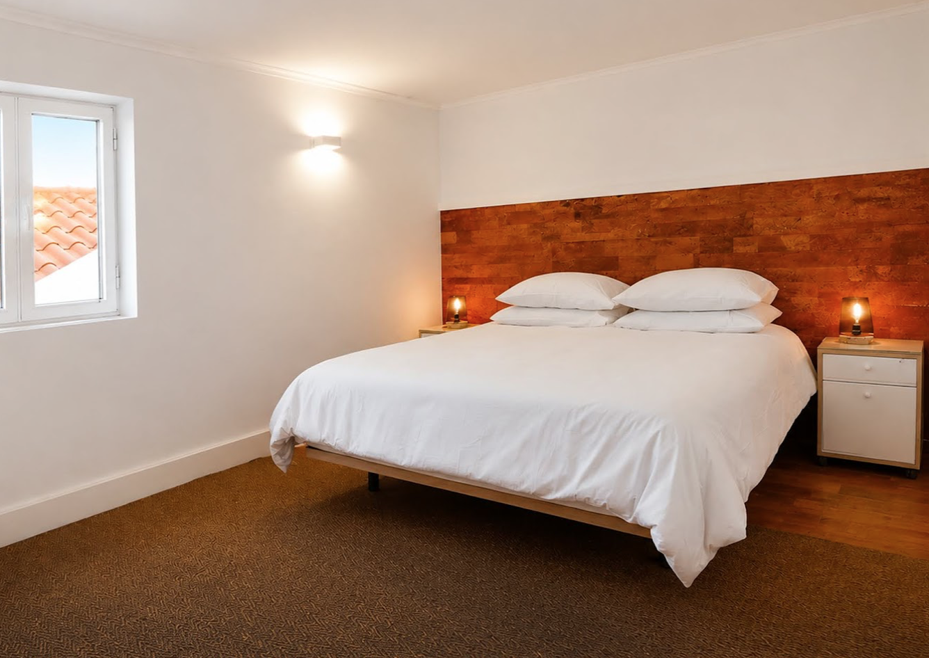 A bedroom with a large bed, white bedding, and multiple pillows. There are two nightstands with lamps on either side of the bed. A small window shows a tiled roof outside.