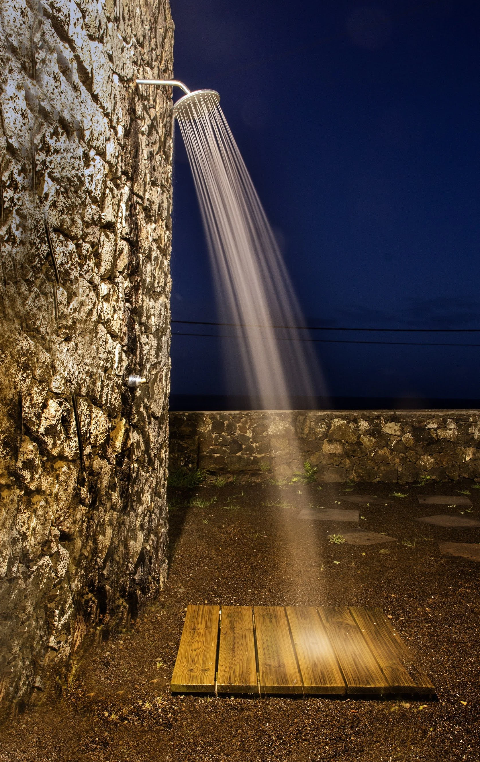 Nighttime outdoor scene with a stone wall, a shower head mounted on the wall, water flowing from it, and a wooden platform on the ground.