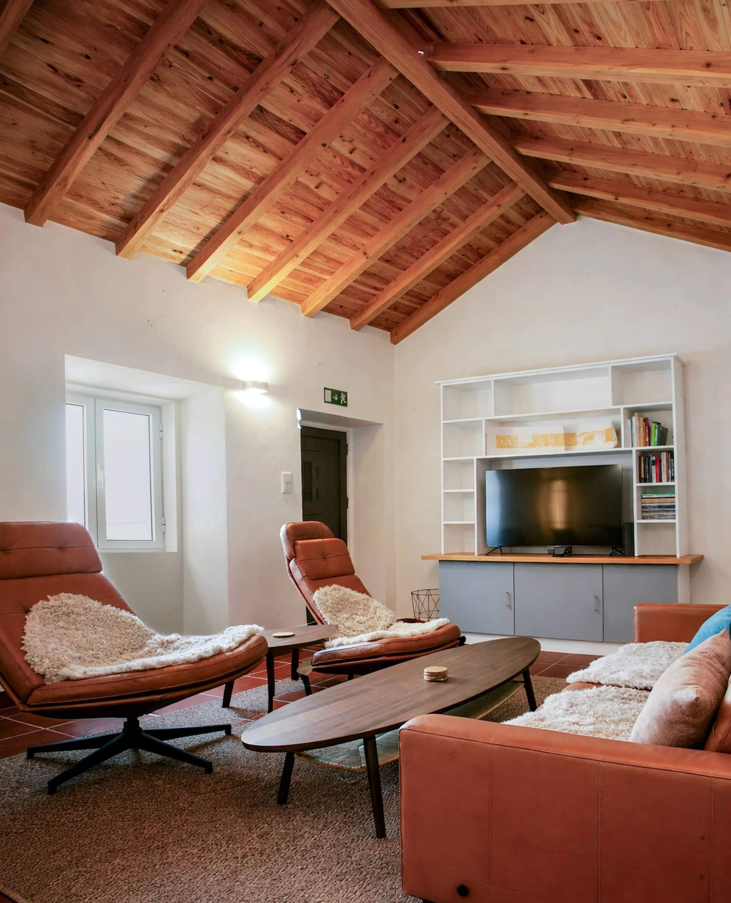 Living room with a wooden vaulted ceiling, a white TV stand with a television, and sofa chairs with plush throws. There is a small window and an exit door, with a rug on the floor.