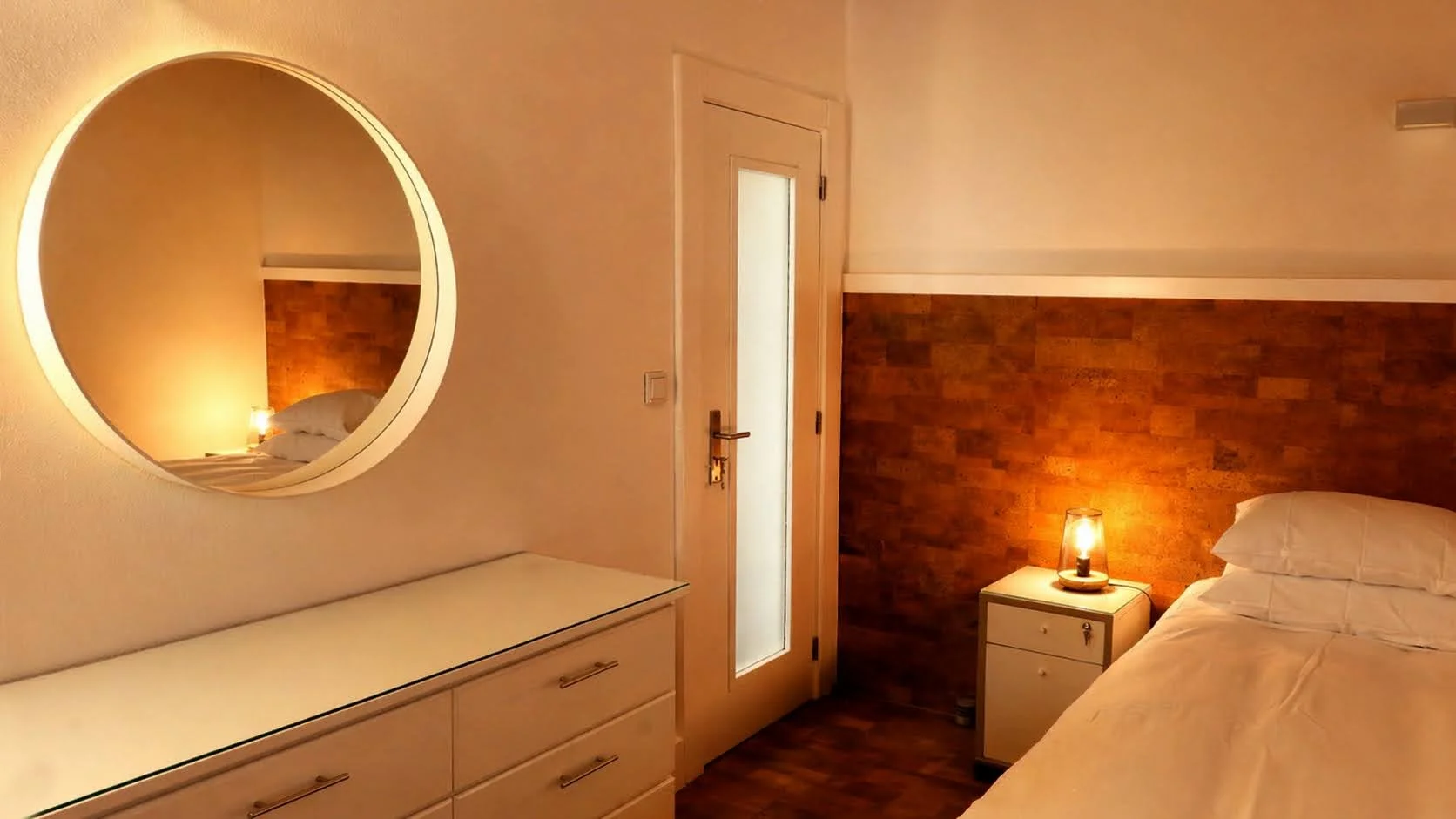 A cozy hotel room with a bed, bedside table, lamp, and a large round mirror reflecting part of the bed and headboard, with warm lighting and wooden accents.