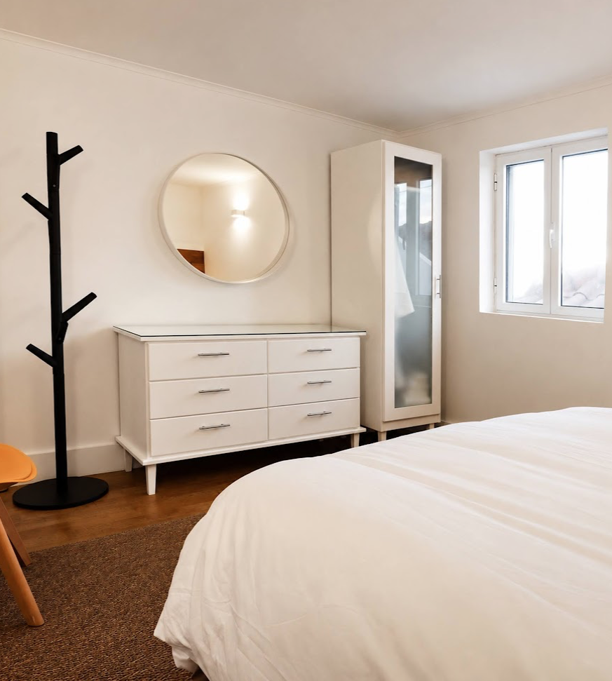 A bedroom corner with a white dresser, a round mirror, a black coat rack, a tall white cabinet with frosted glass door, a window, and part of a bed with white bedding.