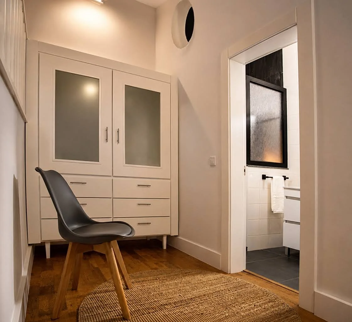 Interior view of a room with a black chair, a white cabinet with frosted glass doors, a woven rug, and a doorway leading to a bathroom with a black-framed window and white tiles.