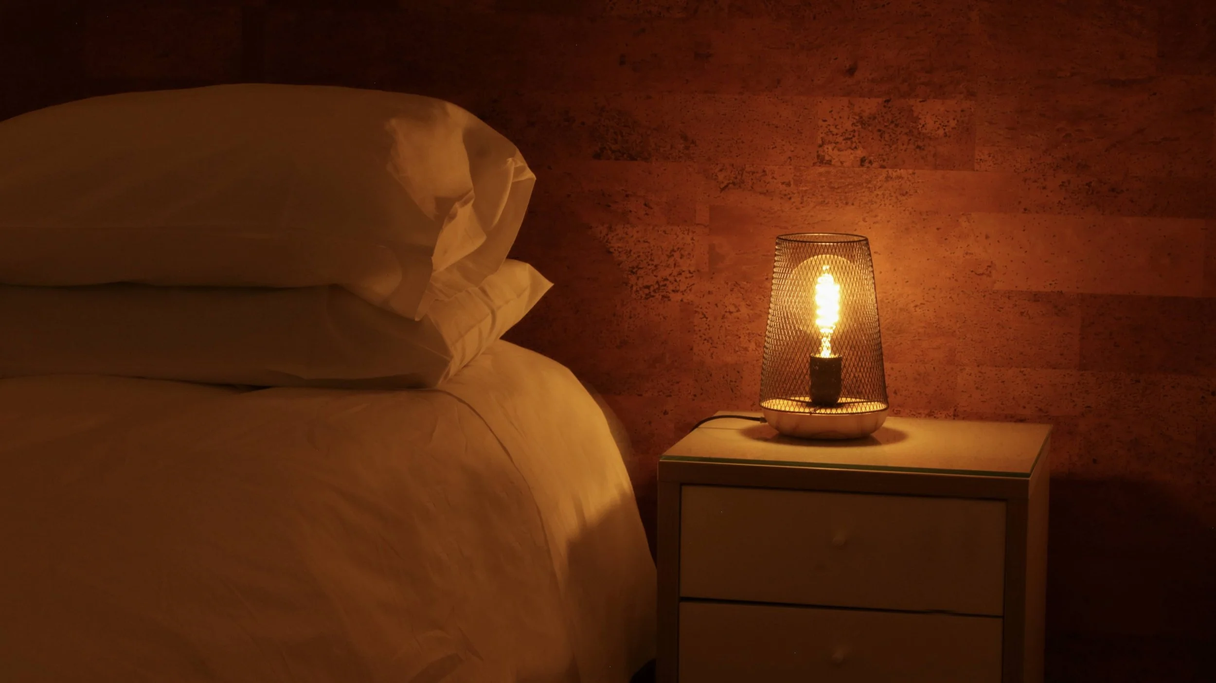 A cozy bedroom corner with a bed featuring white pillows and bedding, next to a nightstand with a modern table lamp emitting a warm glow, against a textured brown wall.
