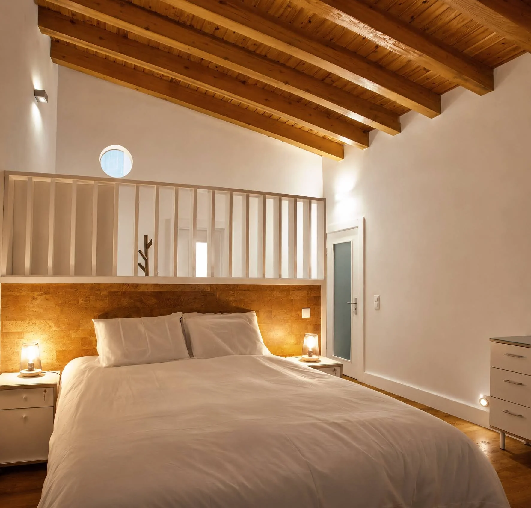 A cozy bedroom with a sloped wooden ceiling, a large bed with white linens, two bedside tables with lamps, and a modern door.