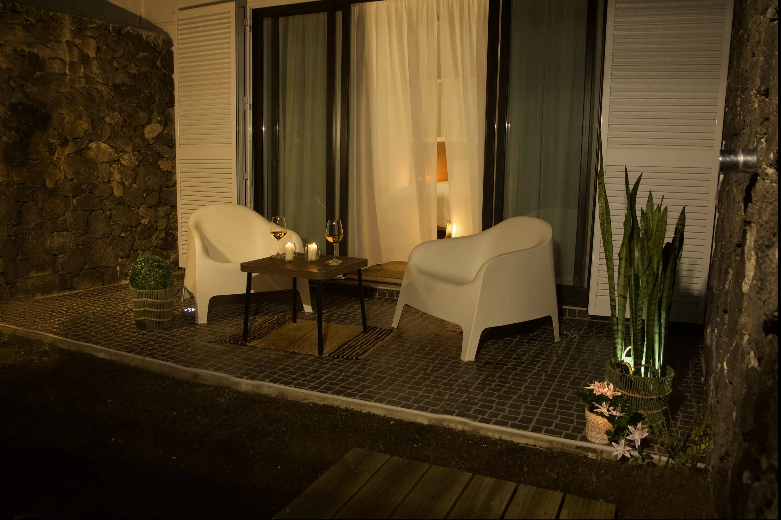 Outdoor patio area at night with two white chairs, a small table holding two glasses of wine and candles, potted plants, and a large window with curtains.