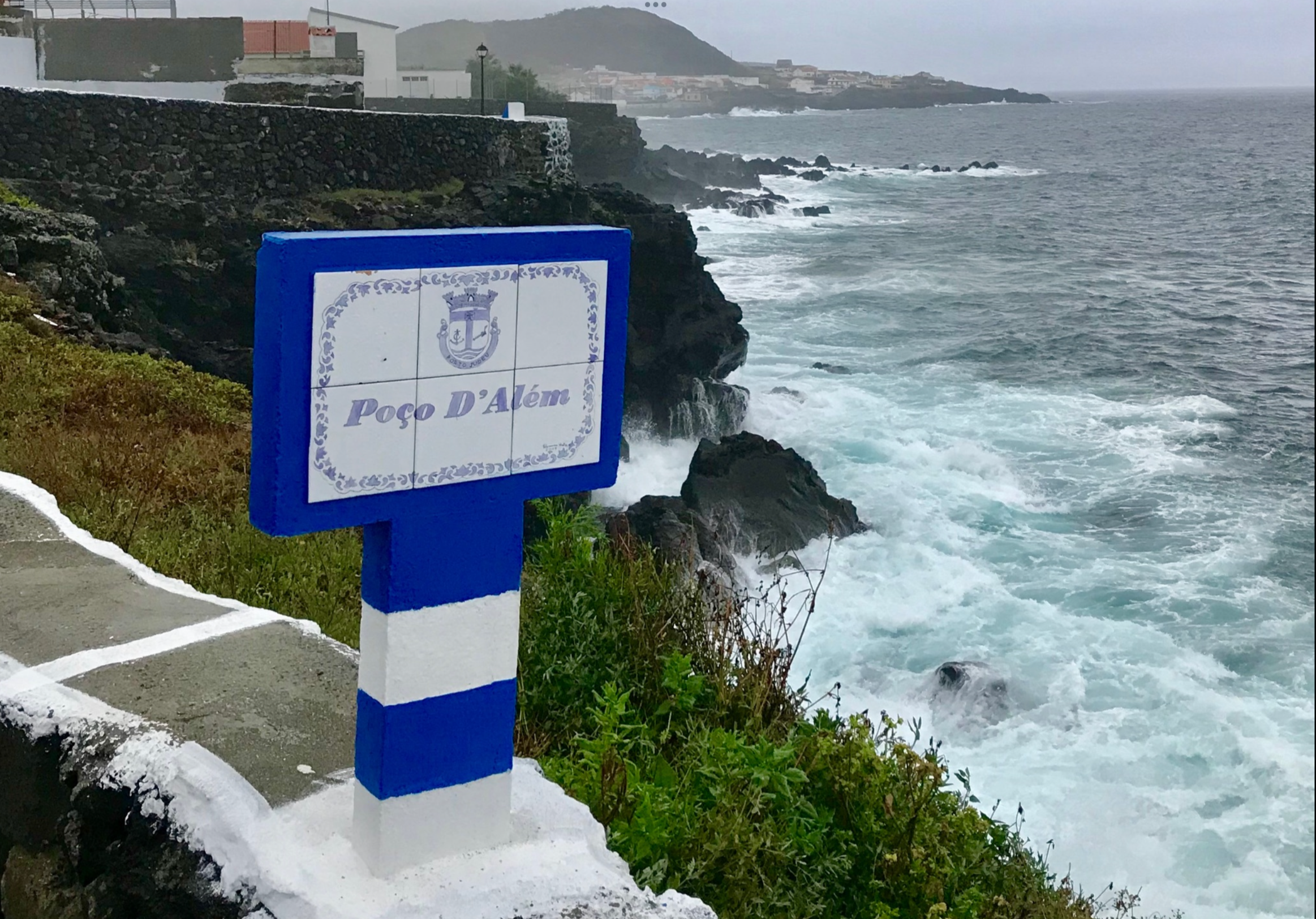 A coastal scene during a rainy day showing a sign with the text 'Poço D'Além' near the rocky shoreline, with waves crashing against the rocks and residential buildings with white and red walls on a hillside in the background.