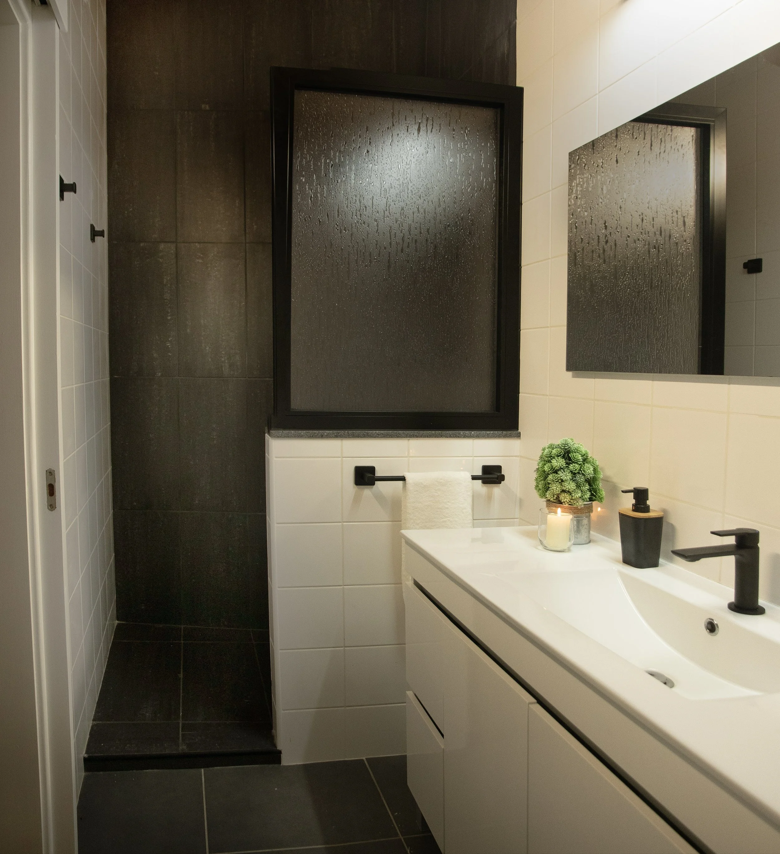 A modern bathroom with black and white tiles. Two frosted glass bathroom partitions with black frames are visible, with steam or water droplets on the glass. The white vanity counter has a black faucet, a black soap dispenser, and a candle. There is 