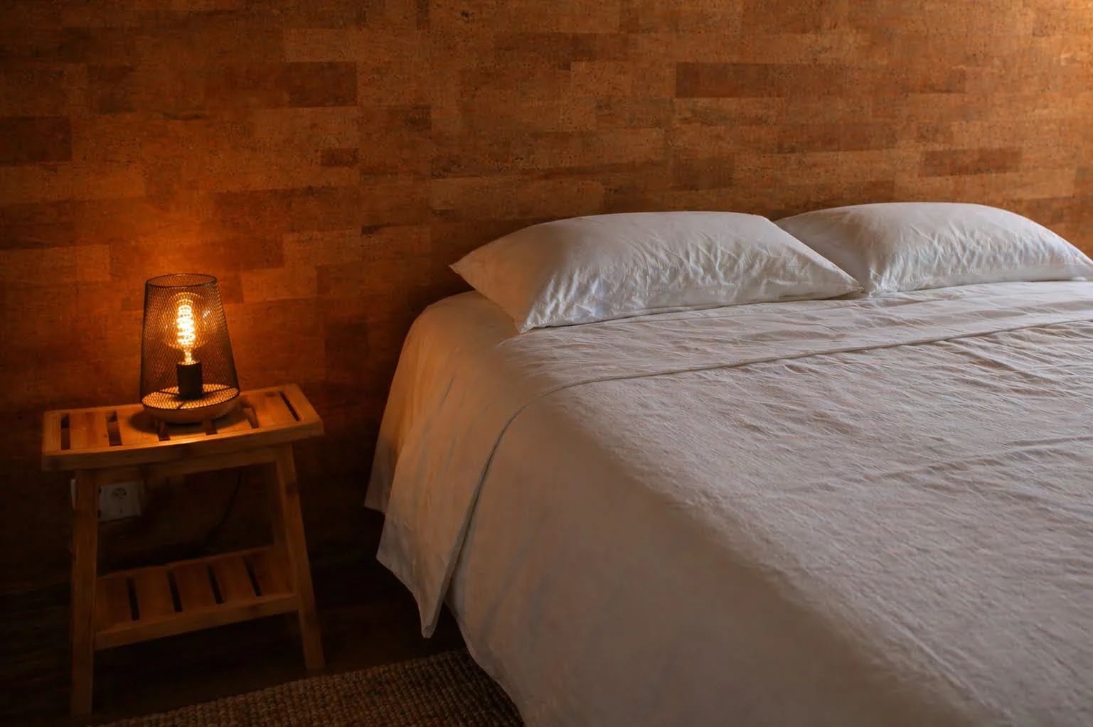 A neatly made bed with white sheets and two pillows, against a wooden-paneled wall, with a small wooden bedside table holding a glowing Edison-style light bulb lamp.