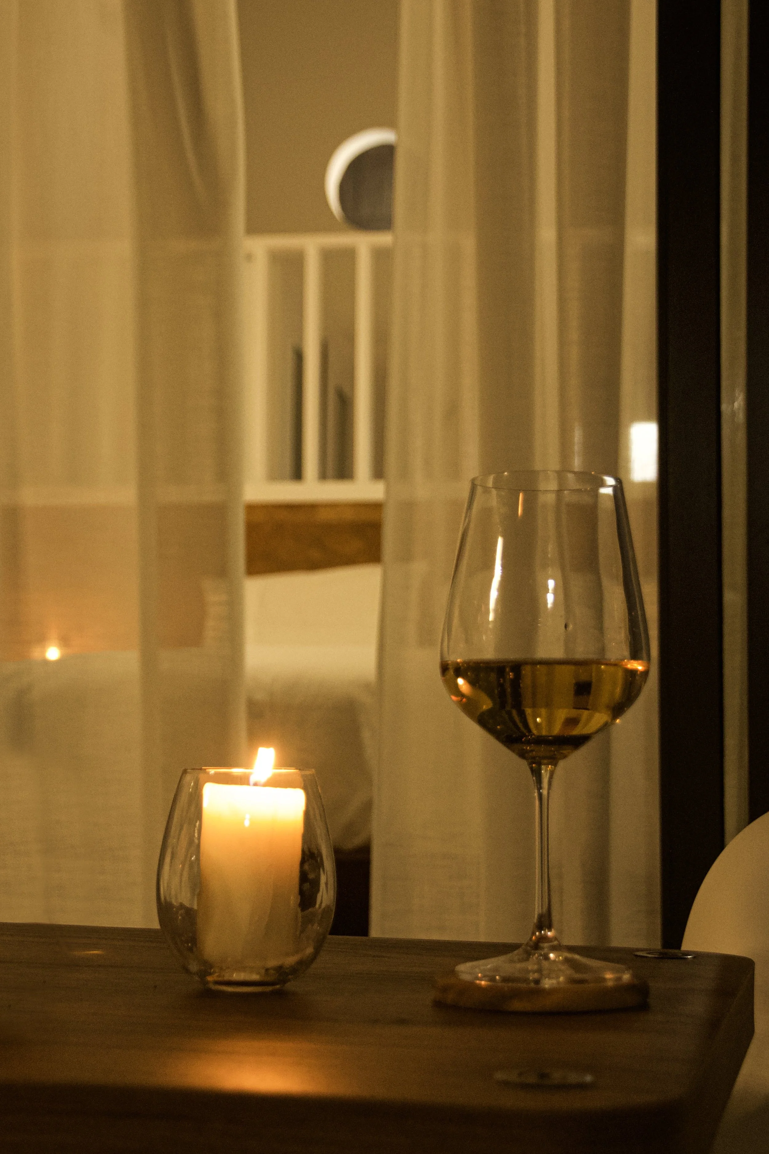 A lit candle in a glass holder and a glass of white wine on a wooden surface, with a window, curtains, and a round window behind them.
