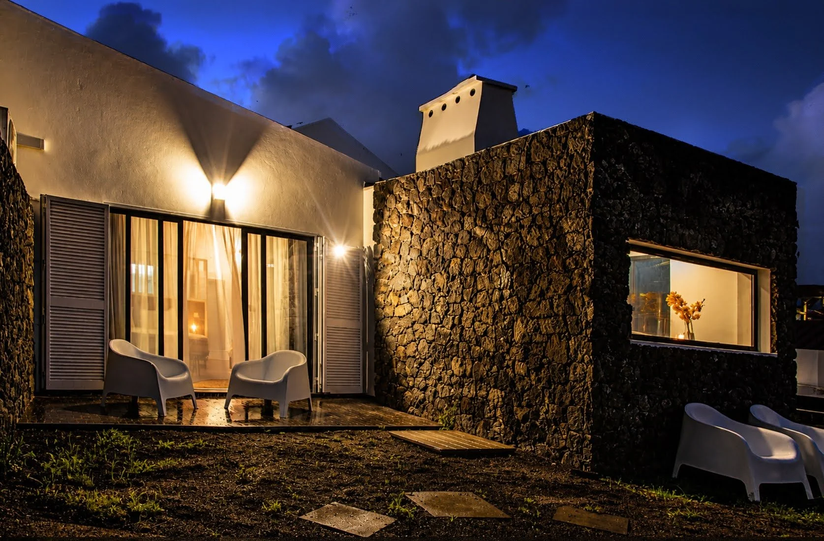 Modern house exterior at night with large glass sliding doors, two white chairs outside, warm lighting inside, and a stone wall on the right side. The sky is dark with clouds.