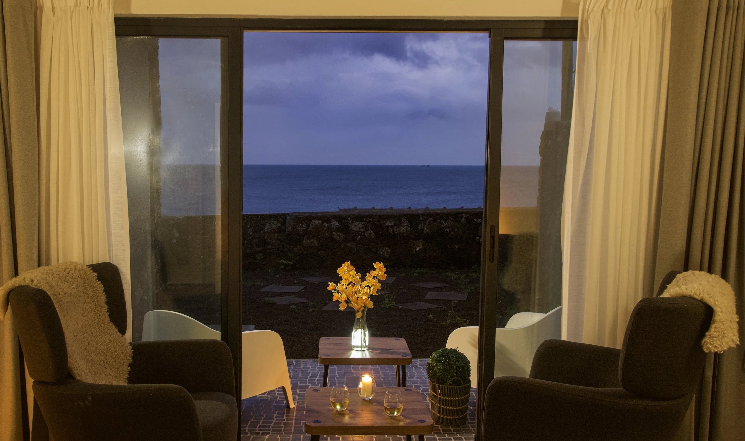 Living room with a sliding glass door opening to a view of the ocean, cloudy sky, and horizon. Inside, there is a small table with a vase of yellow flowers and candles, and surrounding chairs with cozy throws.