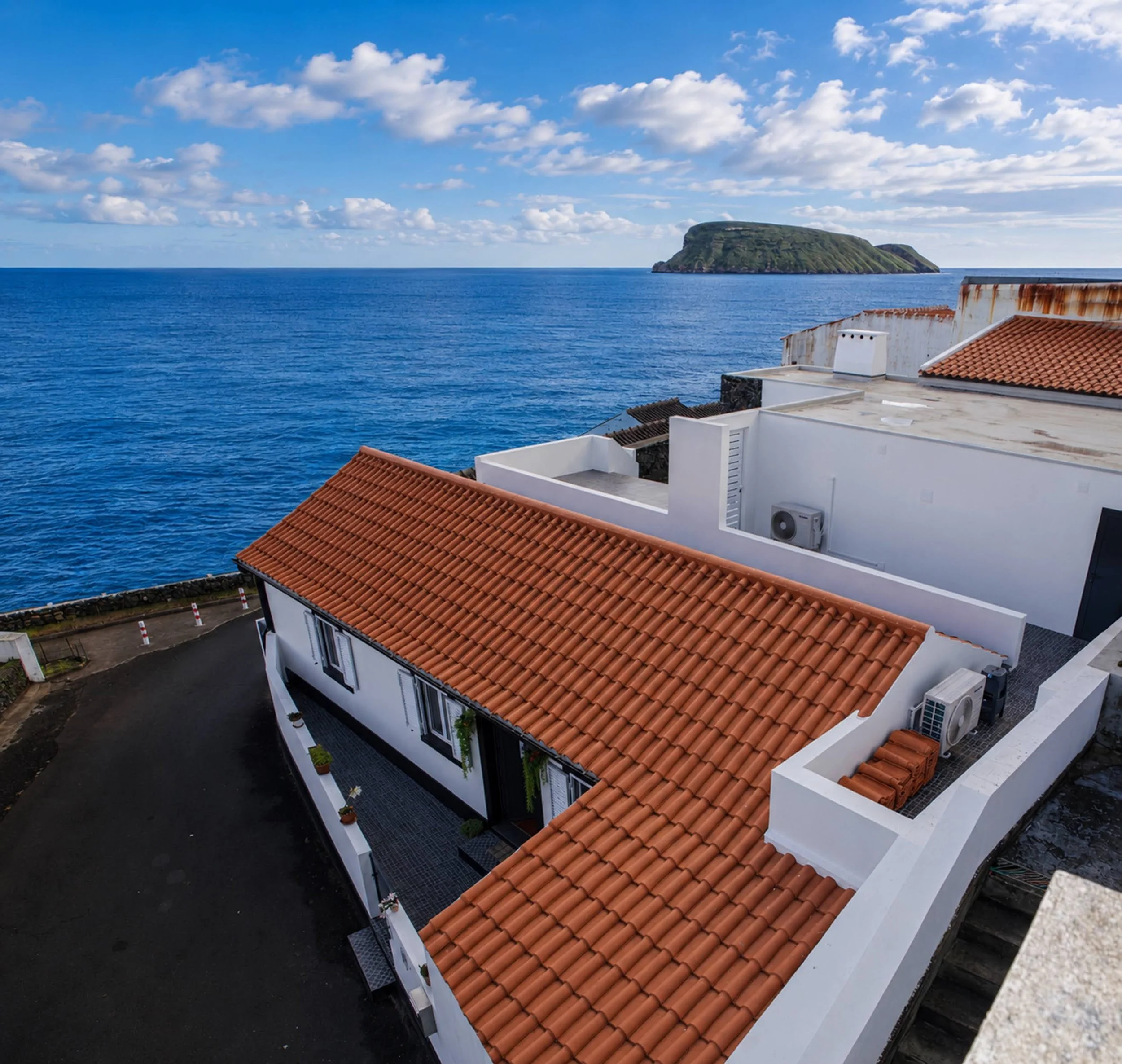 A coastal view showing the sea, a small island with green land, and rooftops of white buildings with red tiles in the foreground.