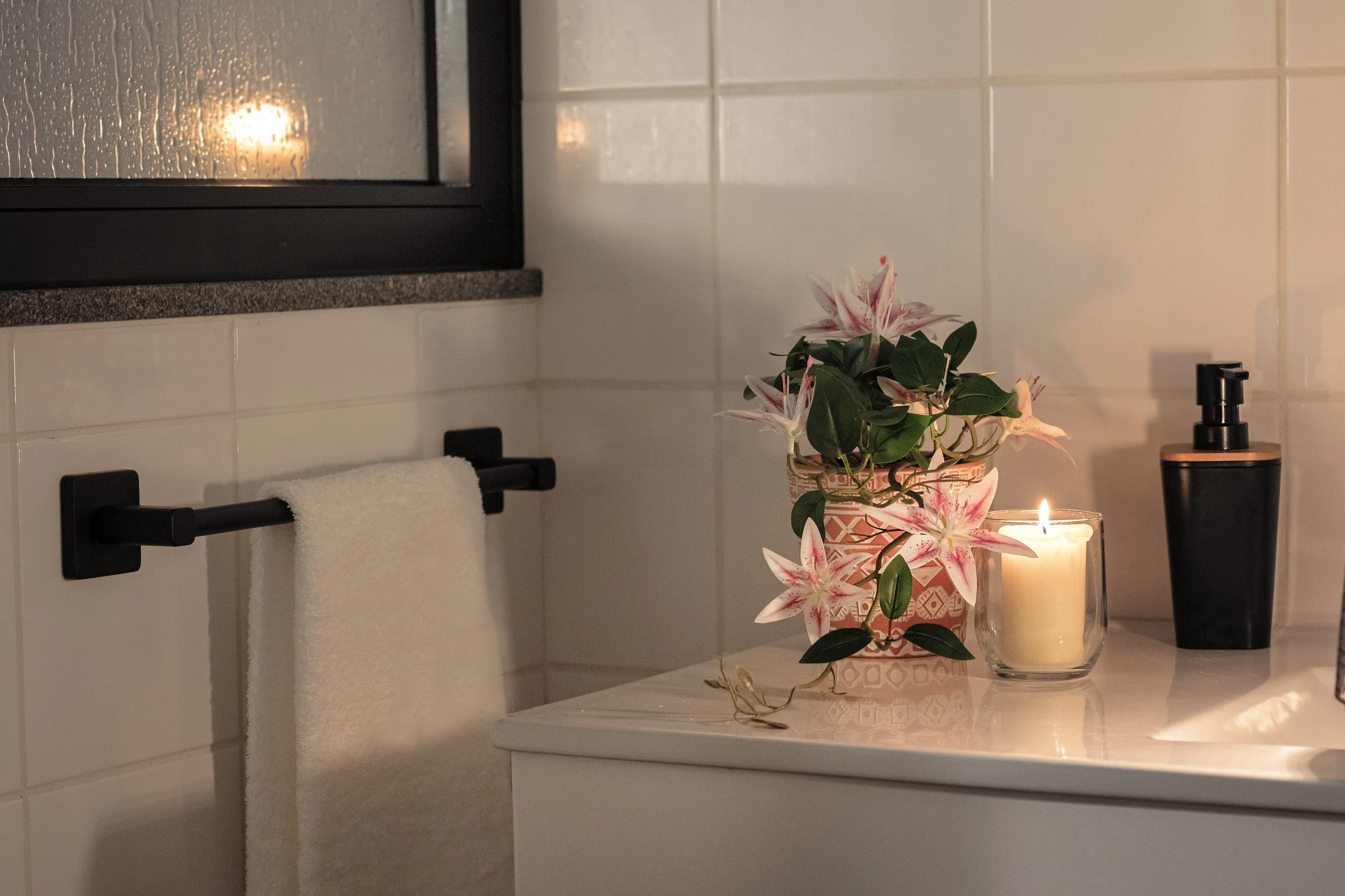 A bathroom countertop decorated with pink and white flowers in a patterned vase, a lit candle, a black soap dispenser, a black towel holder with a white towel, a soap dish, a soap pump, and a window with rain on the glass.