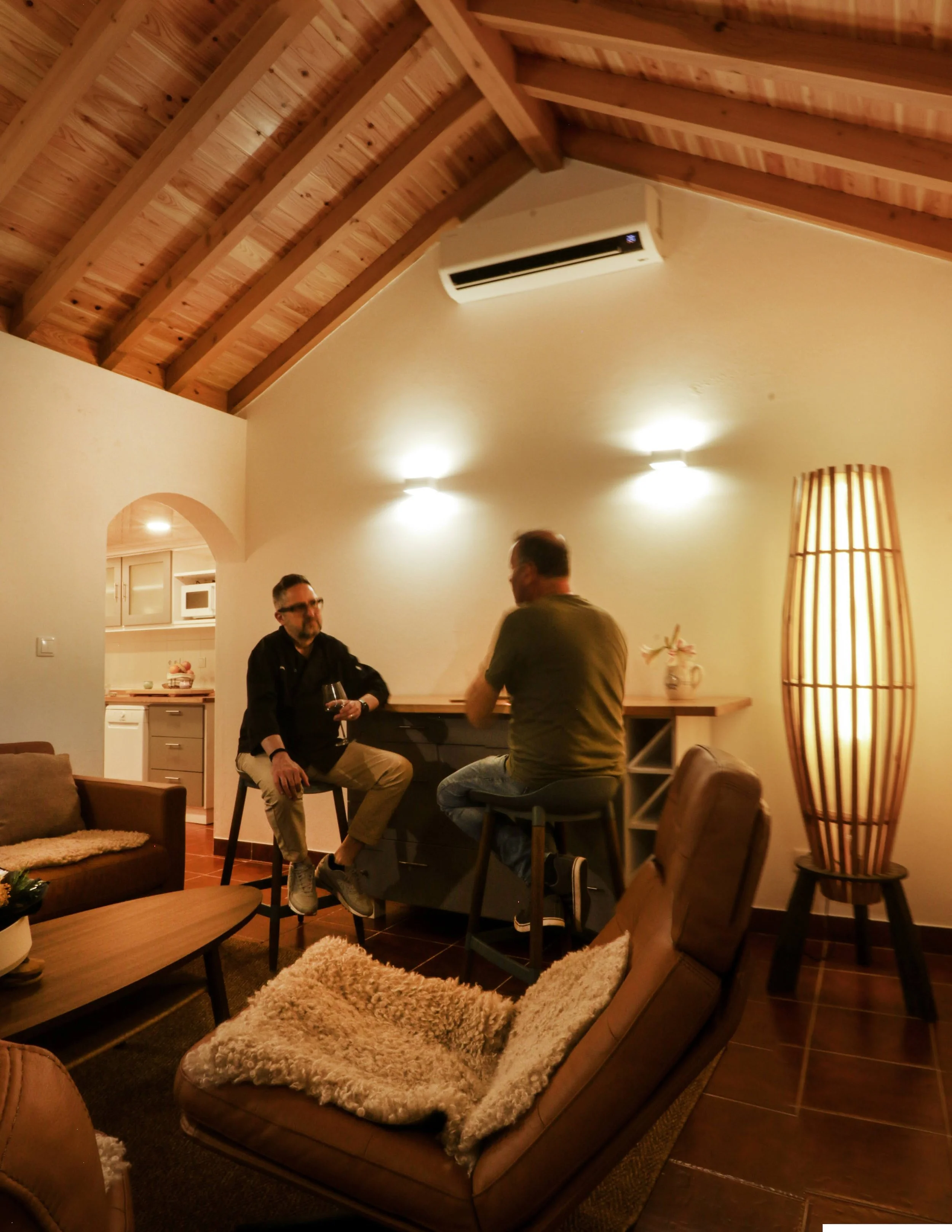 Living room with a wooden ceiling, beige walls, a sofa, a wooden coffee table, and people talking near a bar area. There is a tall, wooden floor lamp, a vase with flowers, and an air conditioning unit on the wall.