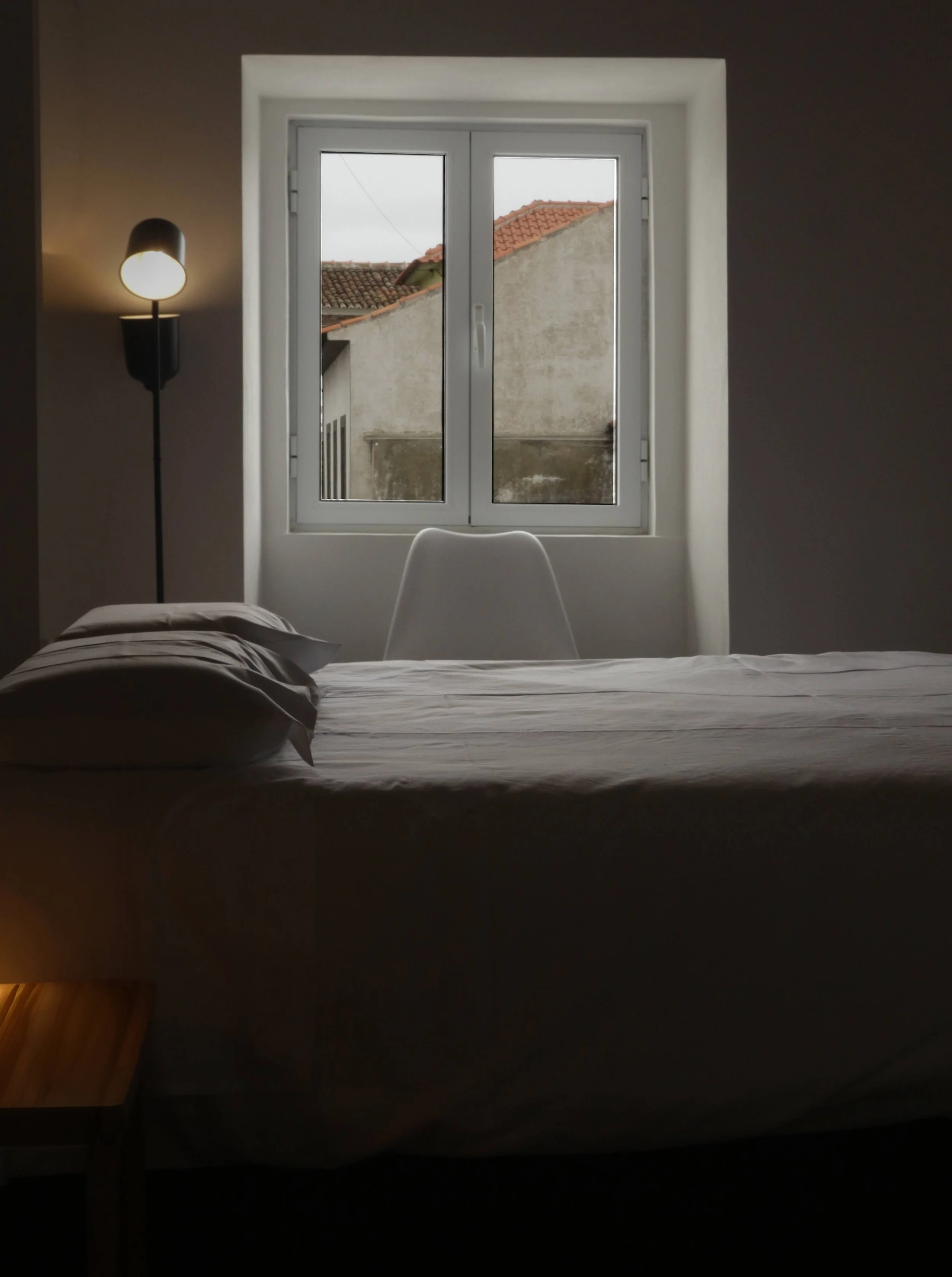 Interior of a bedroom with a bed, pillows, a window showing rooftops outside, and a floor lamp next to the bed.